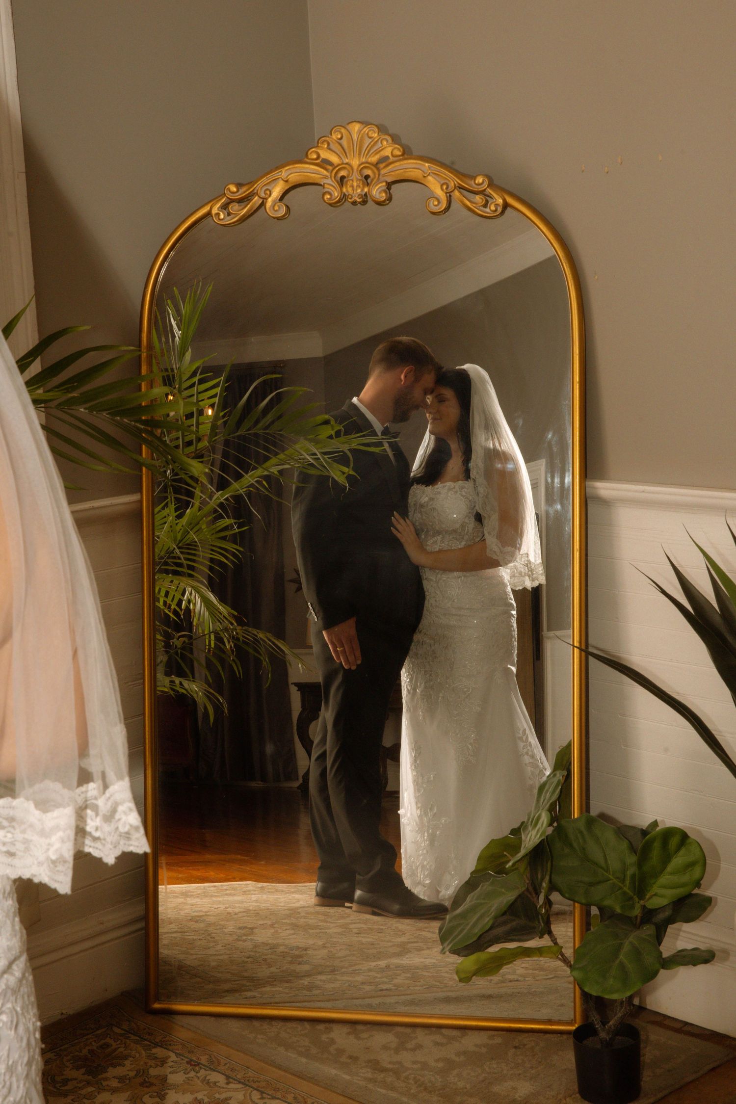 Bride and groom sharing a romantic moment reflected in an ornate gold arch mirror surrounded by lush green plants.