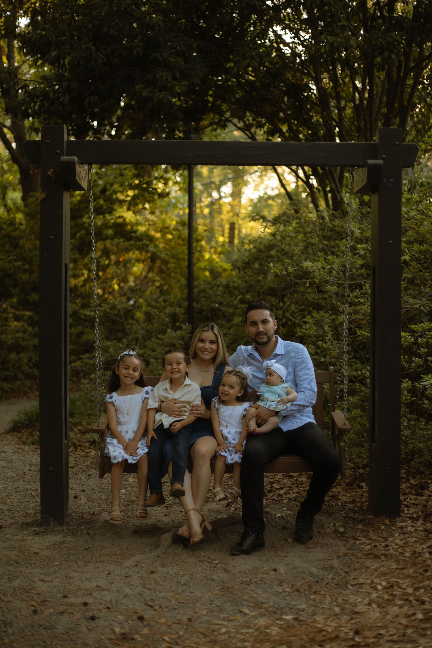 Family of six posing on a wooden swing surrounded by lush green trees in a forest setting.