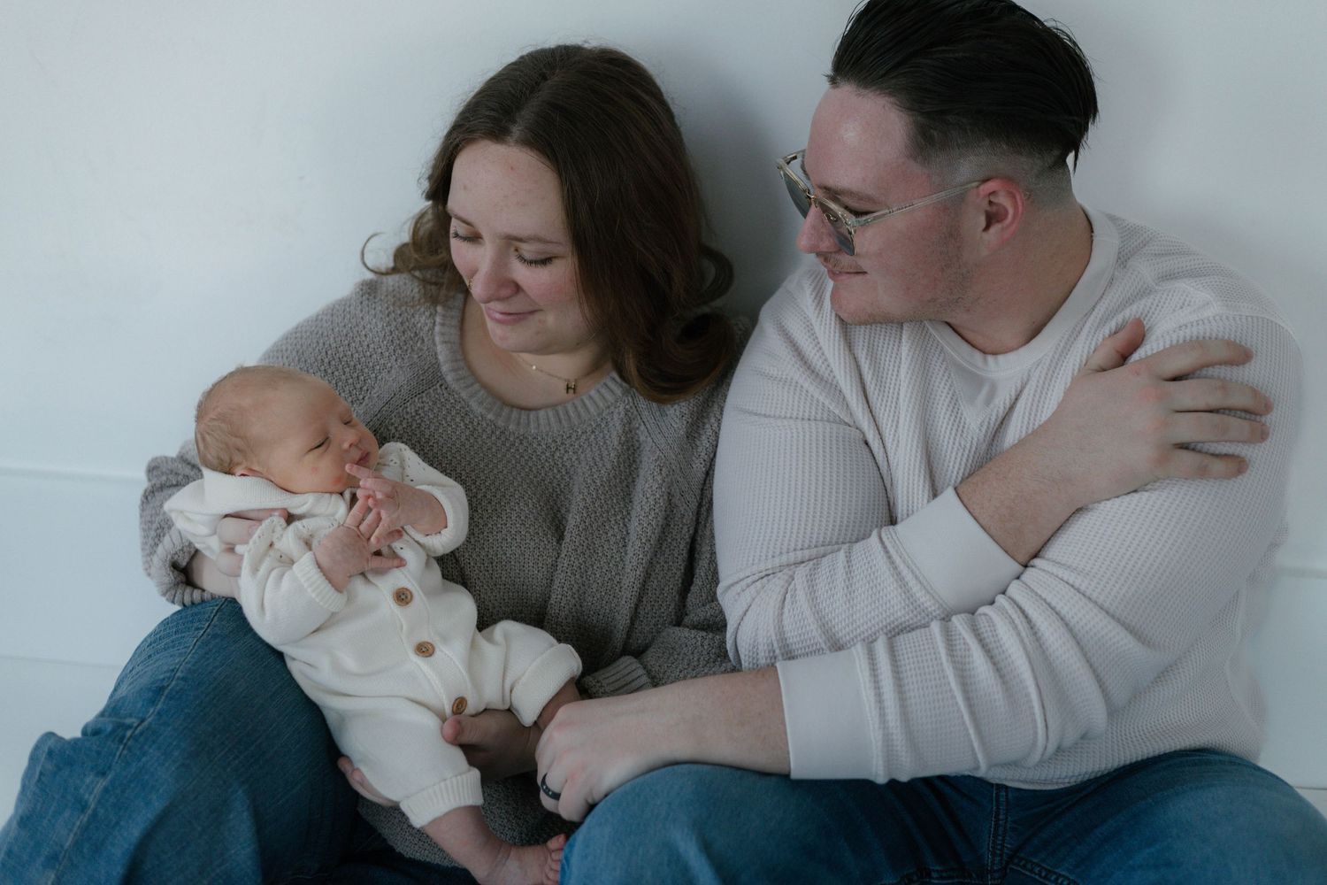 Parents in neutral clothing cuddle with their newborn baby on a cozy bed.
