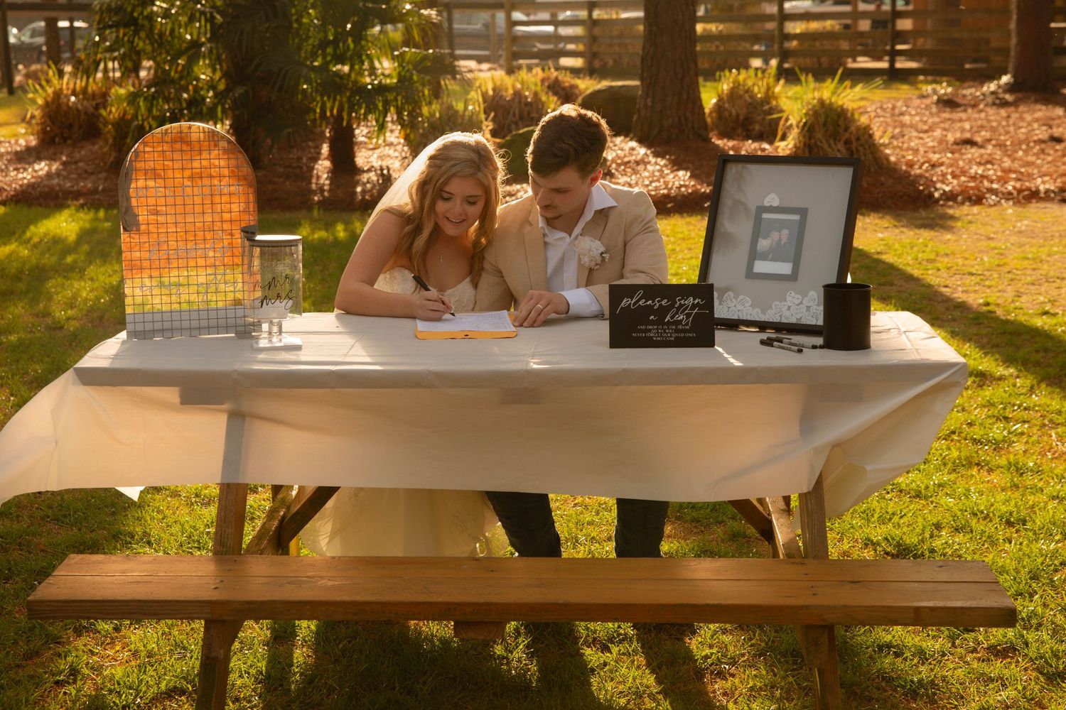 Bride and groom signing wedding registry at white draped table outdoors during golden hour reception.