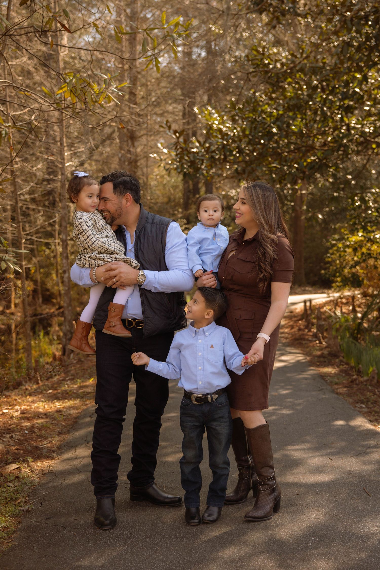 Happy family of five poses on a wooded path in autumn, parents holding young children in coordinated outfits.
