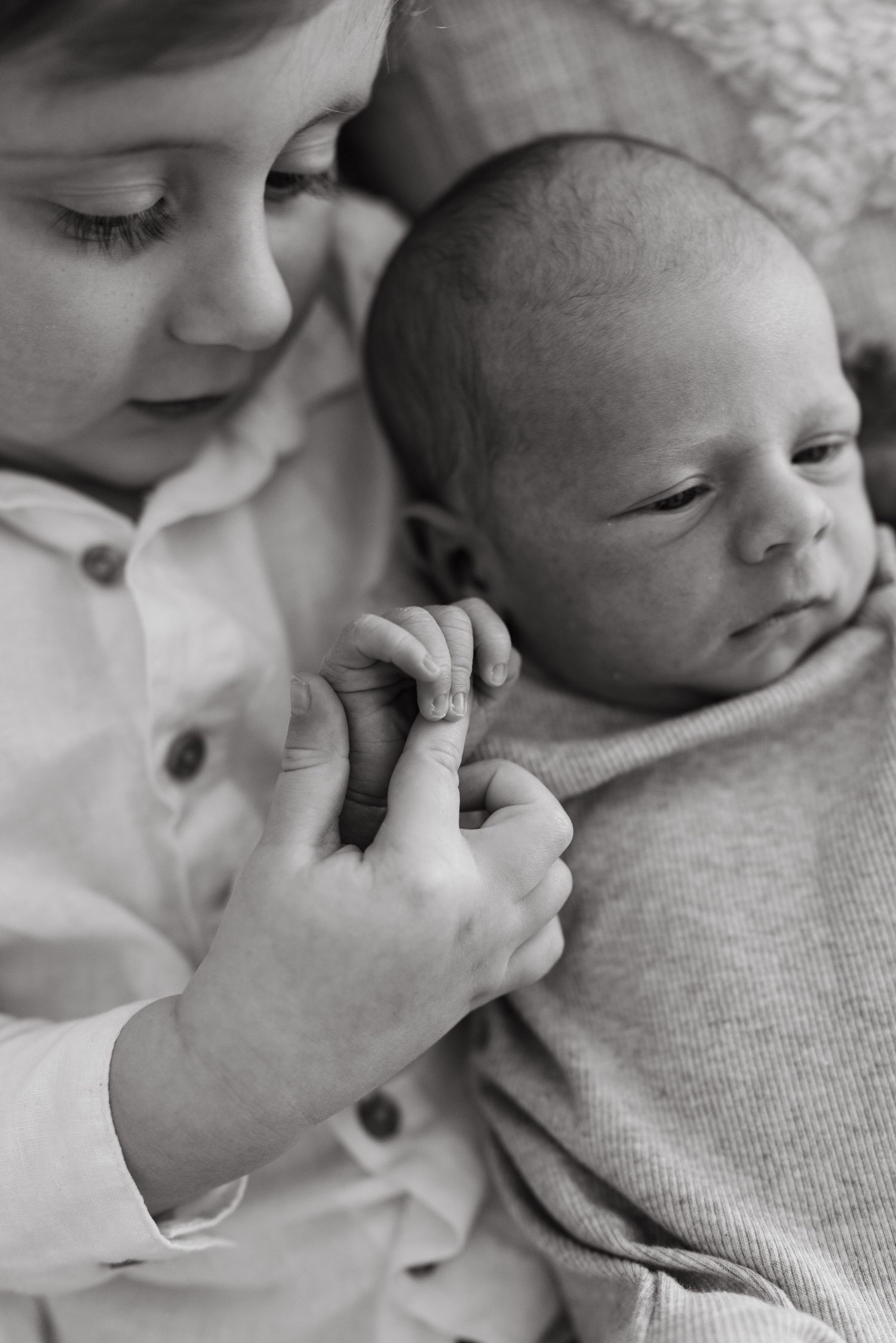 Black and white photo of older child holding newborn baby's hand, a tender sibling bonding moment.