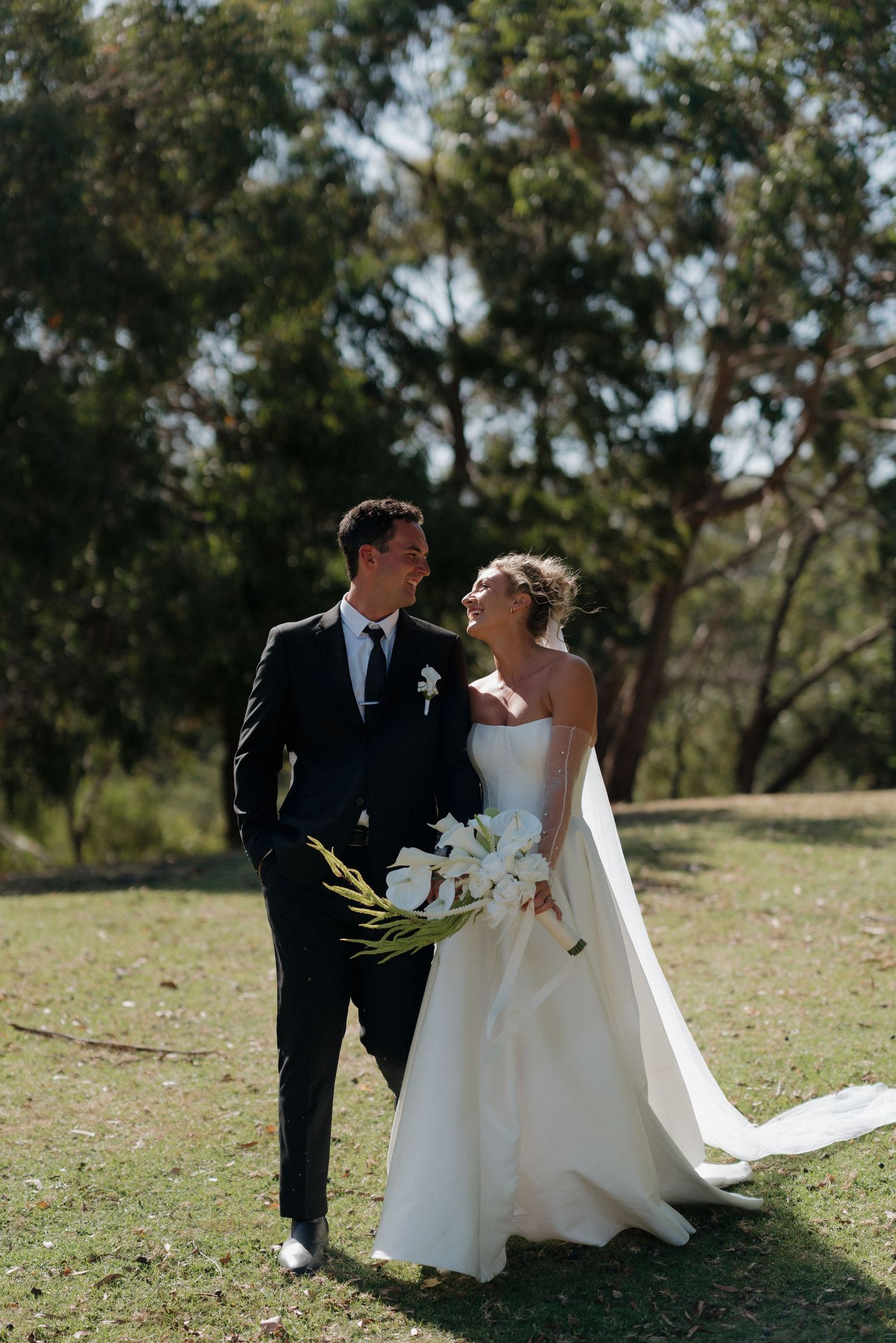 Wedding couple sitting together on grass under sunlit trees.