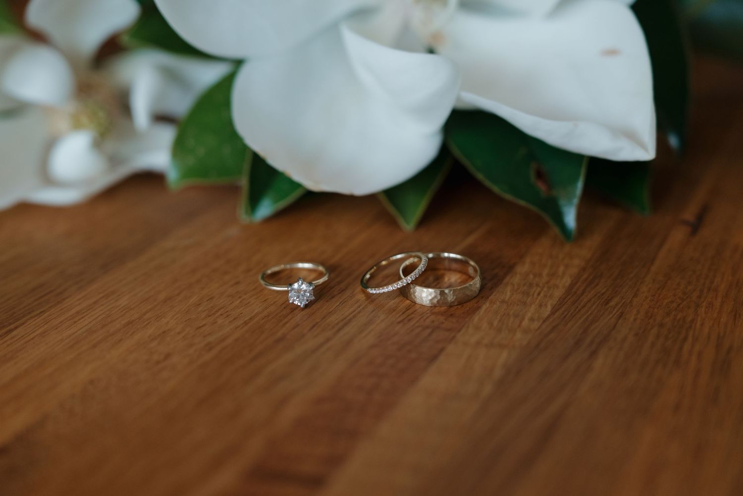 Three gold wedding rings displayed on a wooden surface with white magnolia flowers in the background.
