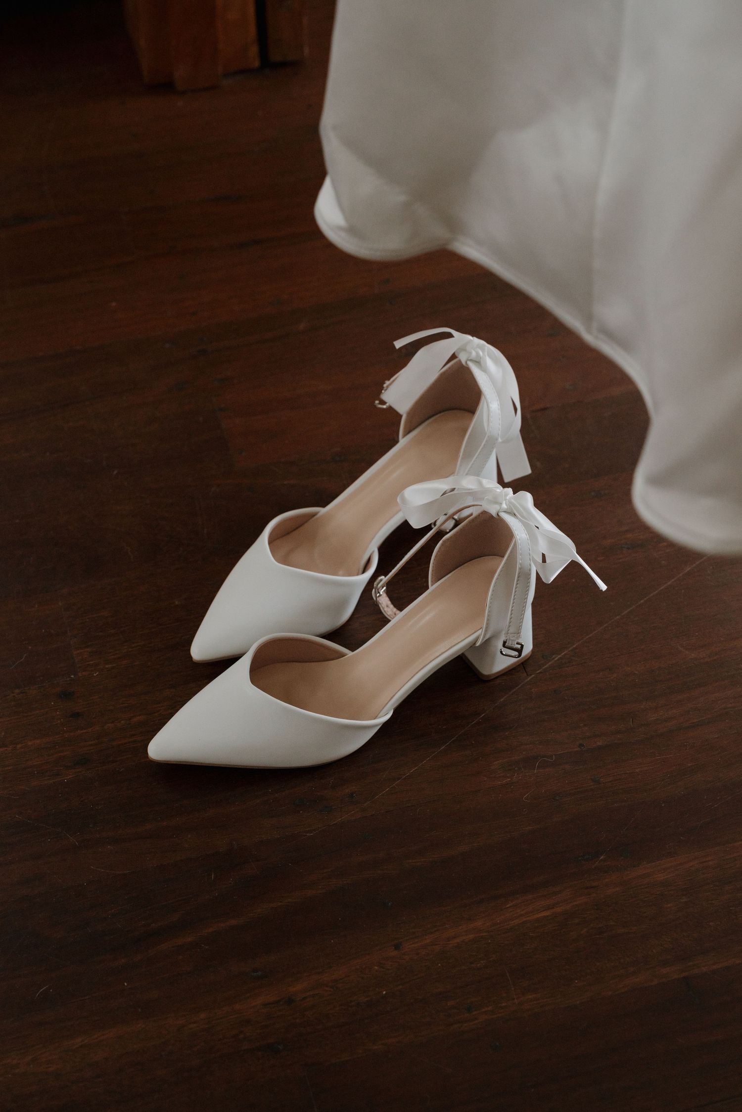 White satin bridal heels with ribbon ankle ties on dark hardwood floor, with wedding dress hem visible in background.