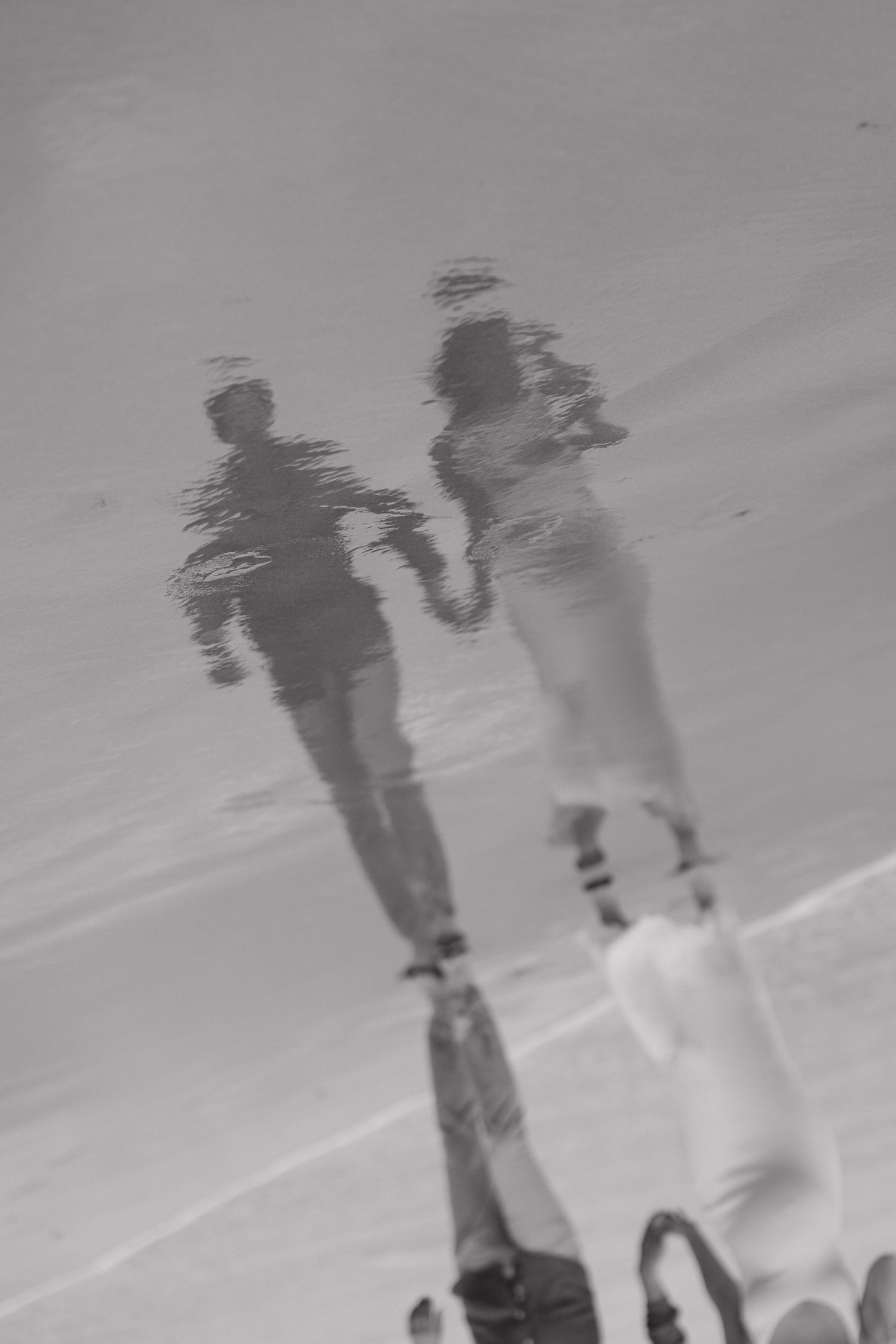 Silhouettes reflected in wet sand on a beach showing two figures walking together.