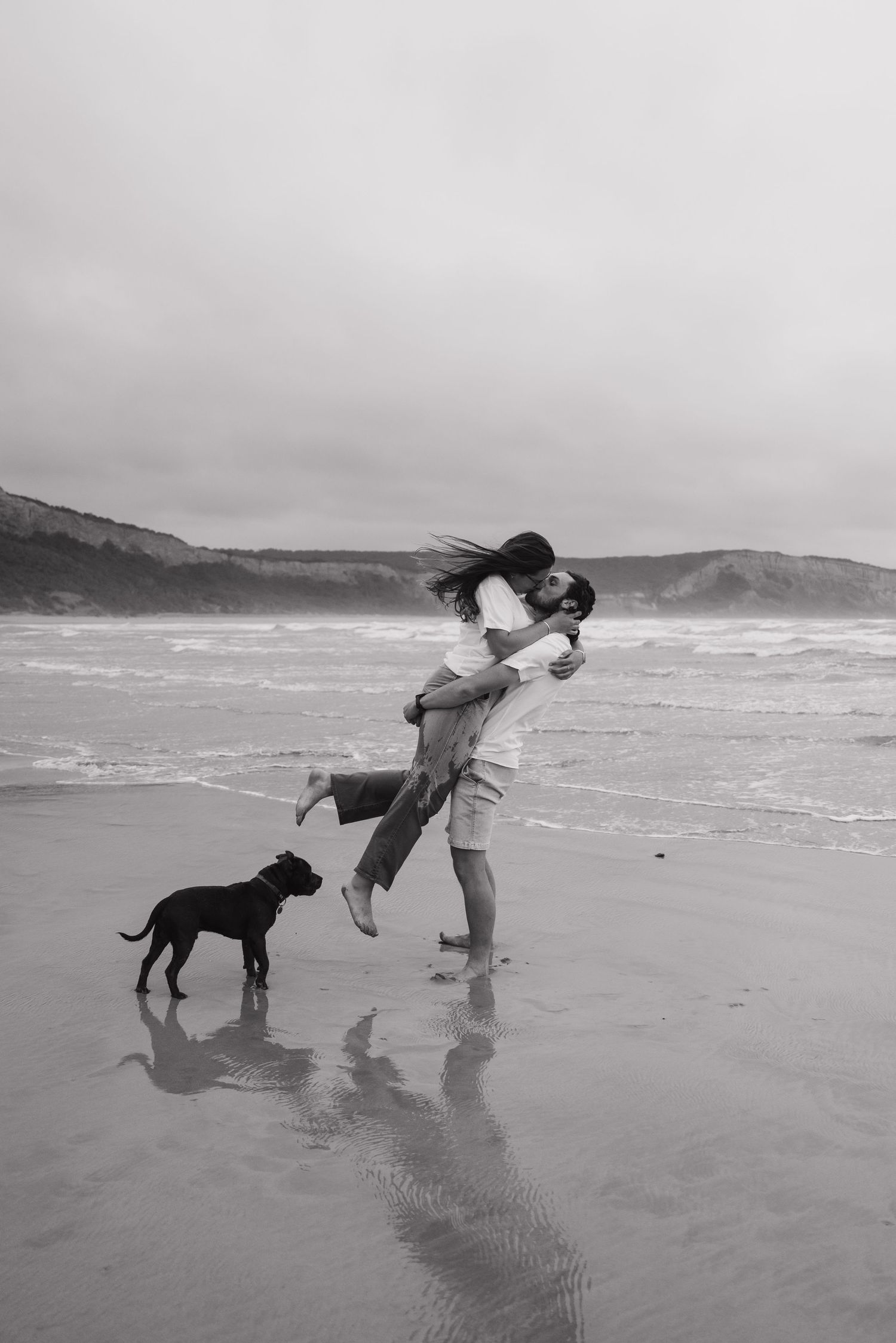 Joyful couple lifting and embracing on a beach with their black dog watching.