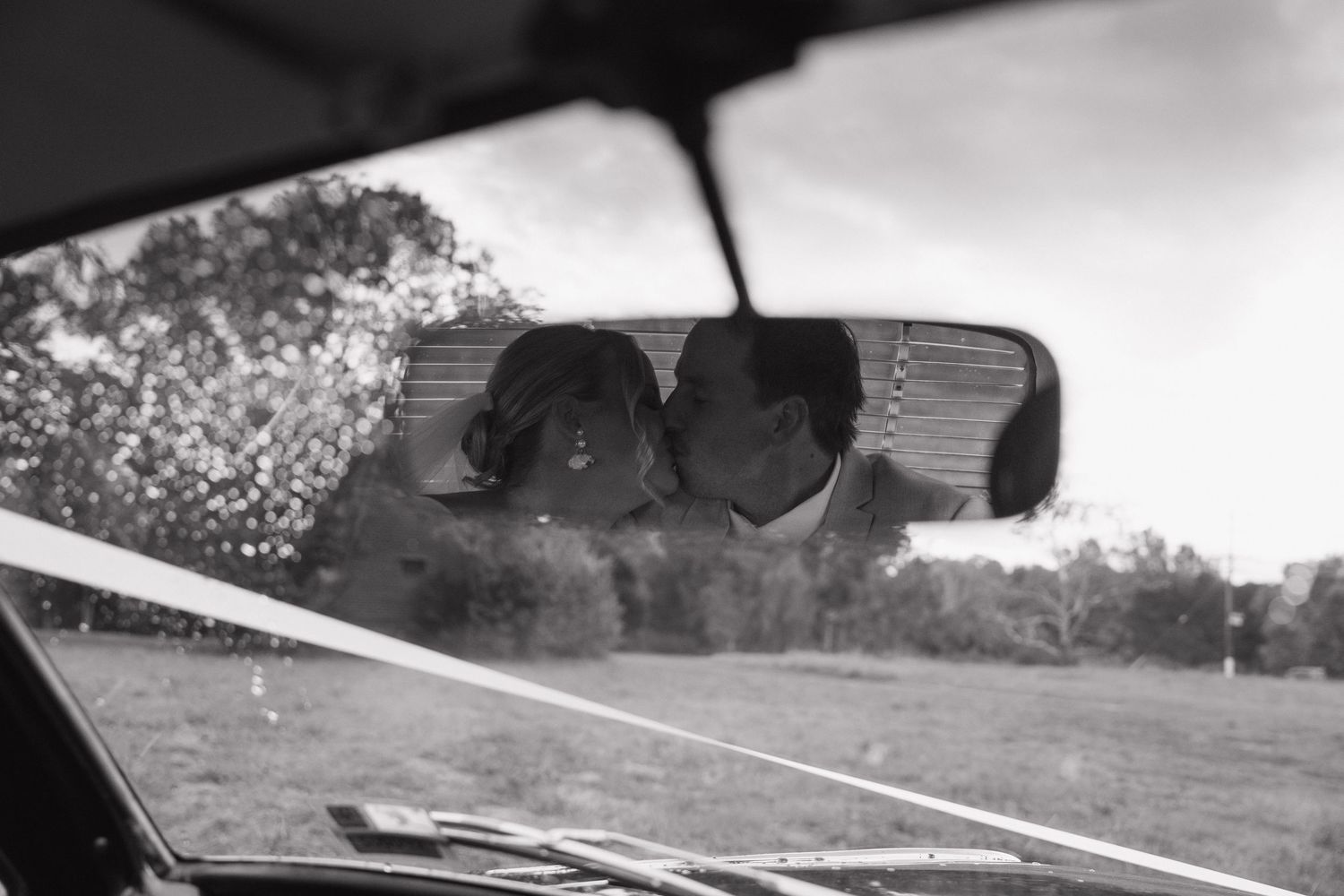 Black and white photo of a couple nearly kissing, reflected in a car rearview mirror with greenery outside.