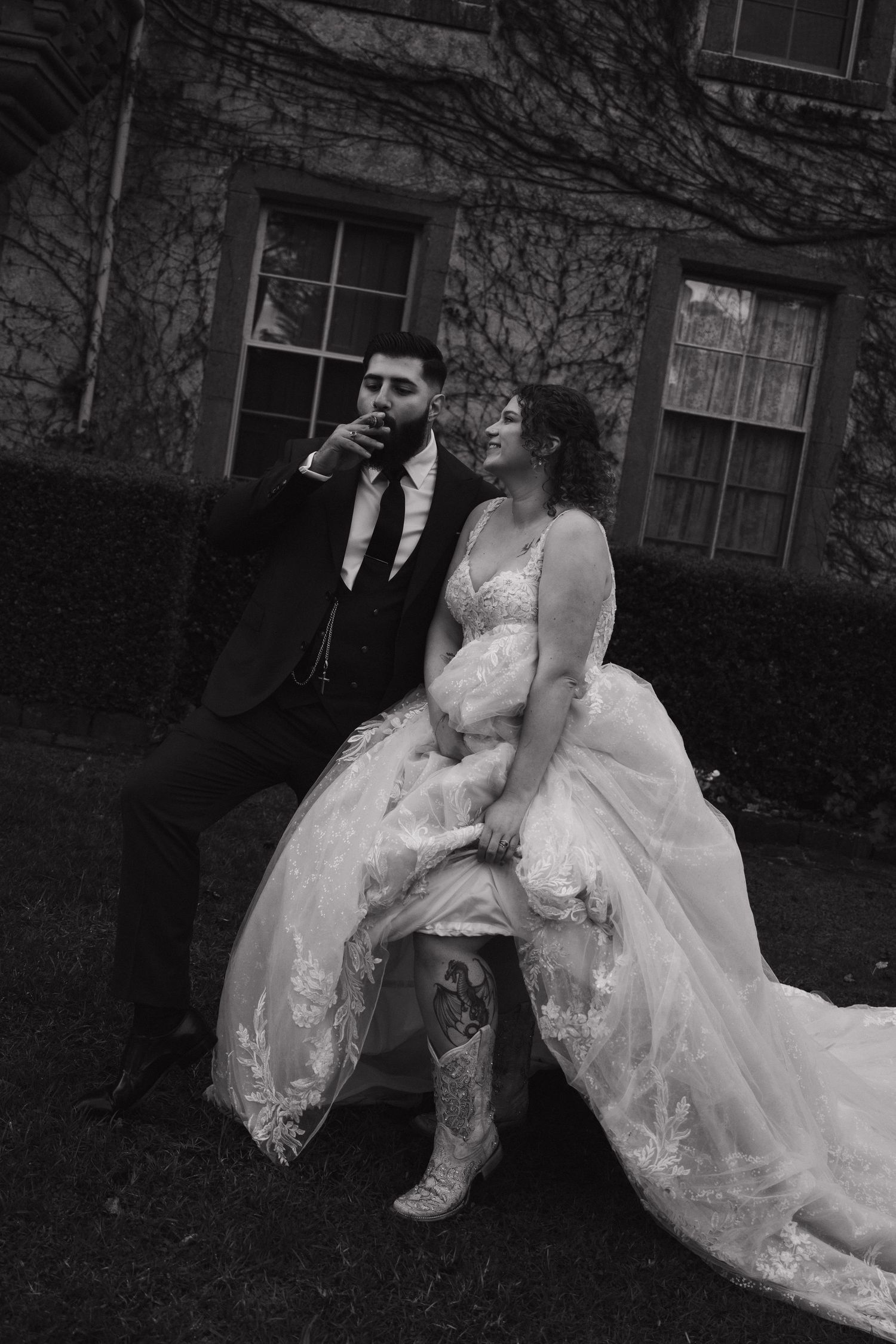 A bride in a white lace wedding dress and boots sits casually outdoors against a stone building backdrop.