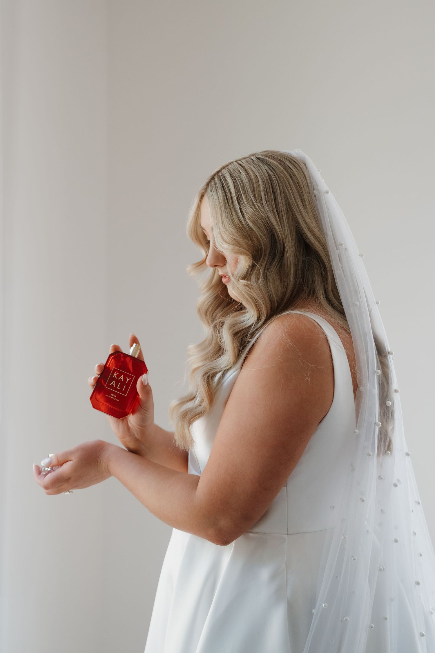 Person in white robe holding red perfume bottle against white wall.