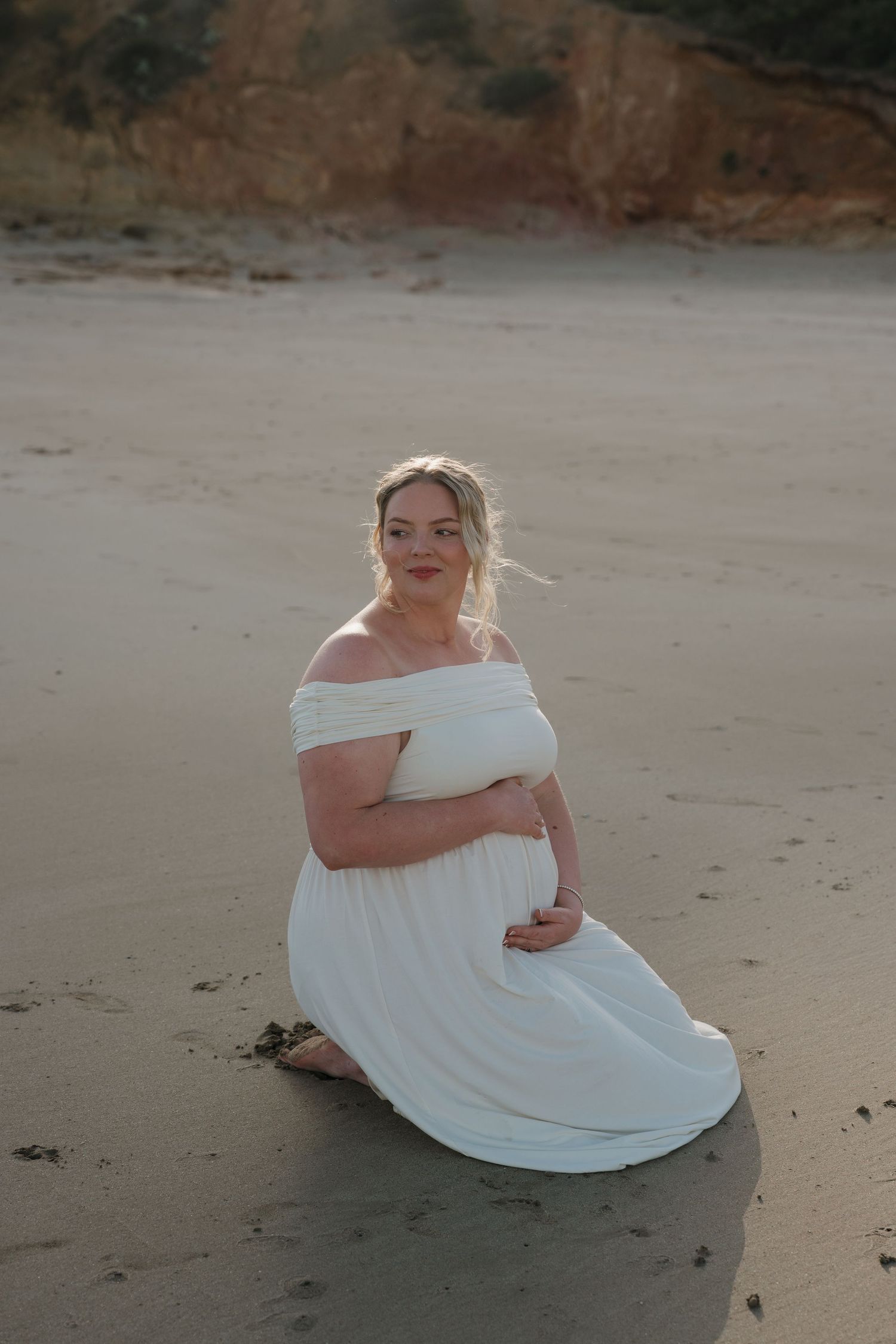 Woman in white dress sitting on beach with cliffs in background.
