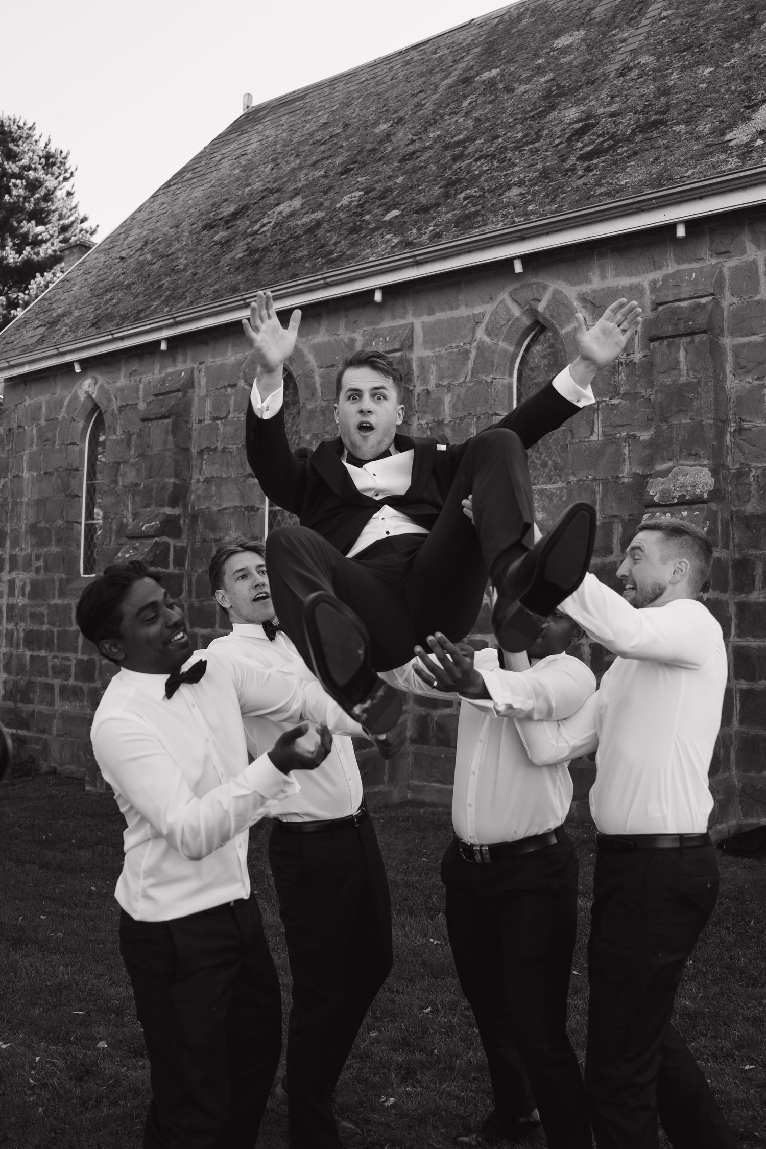 Groomsmen in formal attire toss the groom in the air during wedding celebrations.