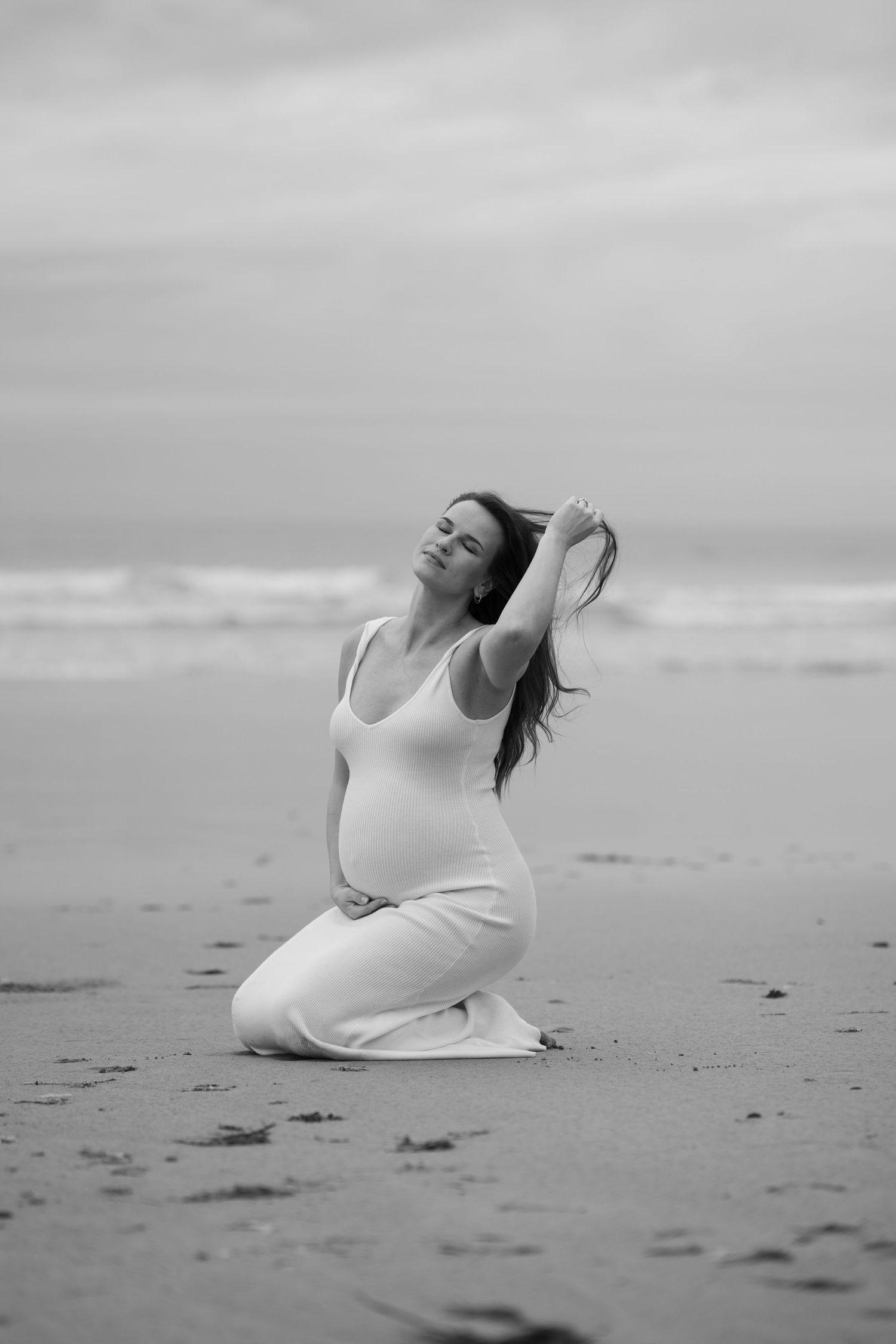 Artistic black and white beach portrait of a woman in white dress sitting in sand.