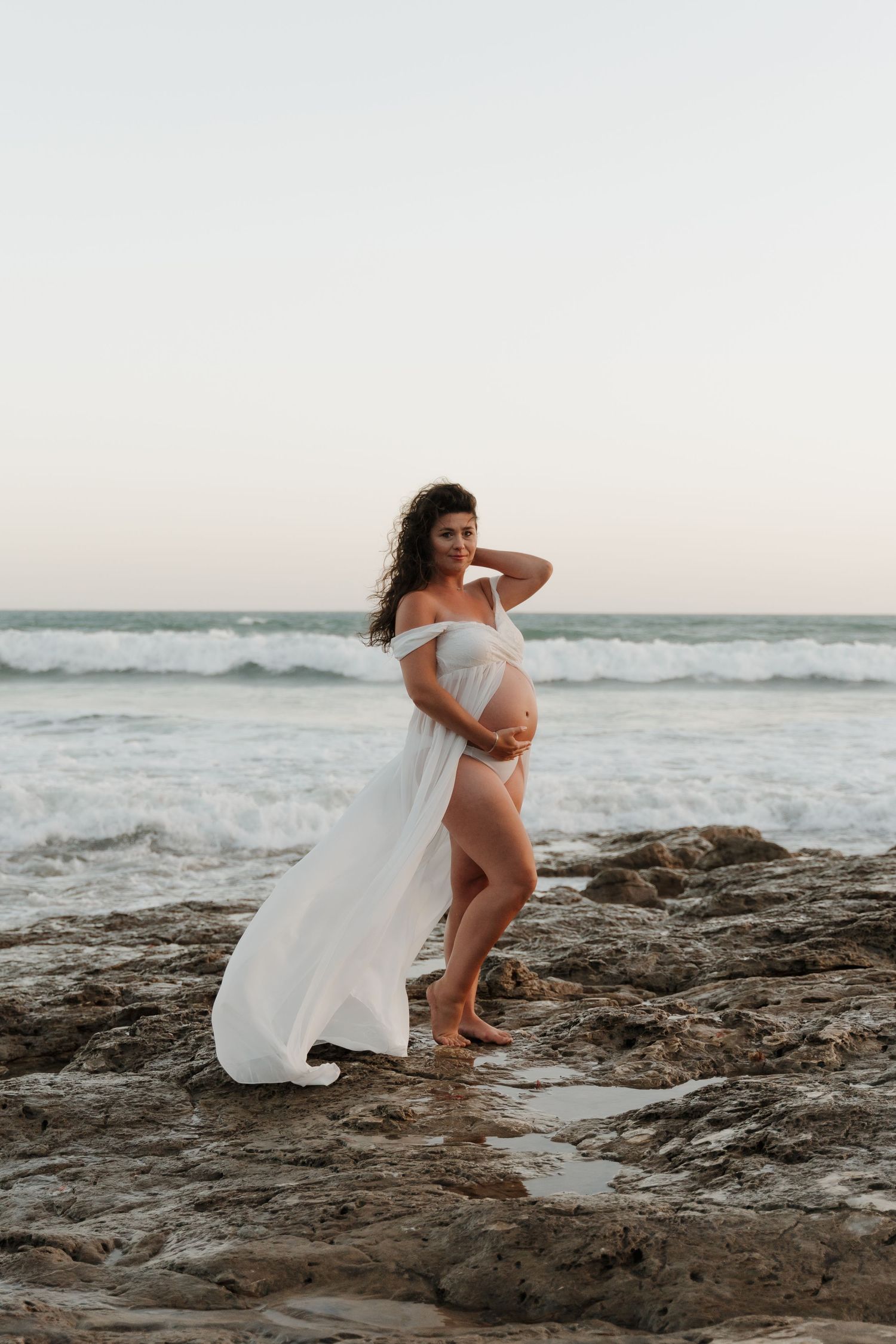 Expectant mother in white fabric poses dramatically on rocky coastal shoreline at sunset.