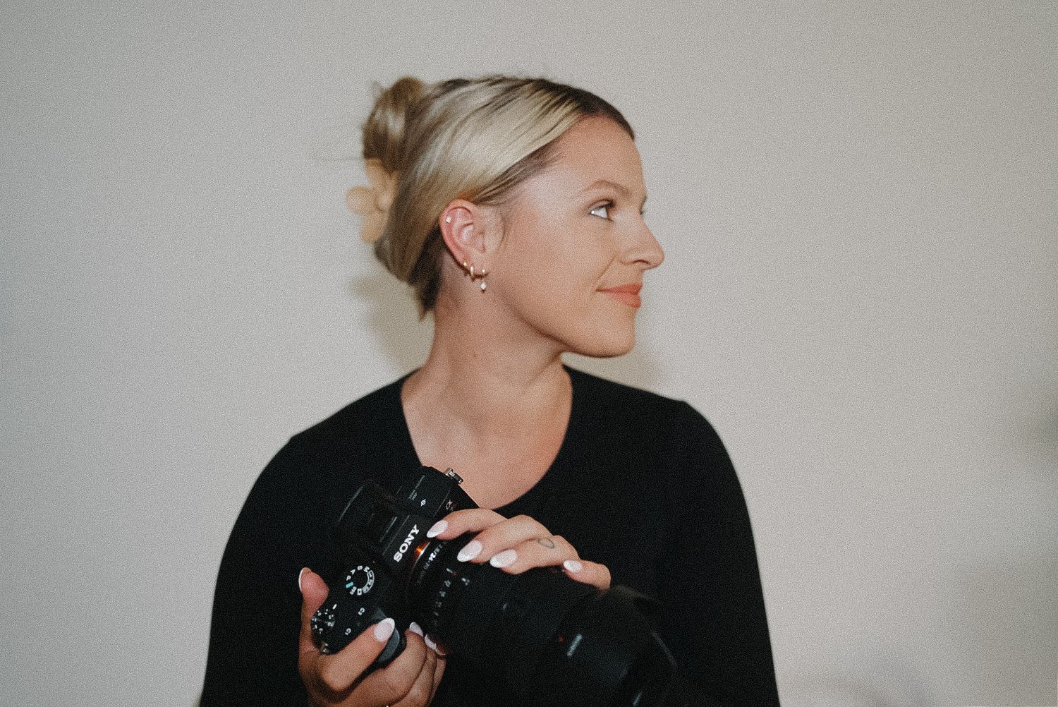 Blonde woman holding a professional DSLR camera, wearing a black top, looking sideways in a bright studio setting.