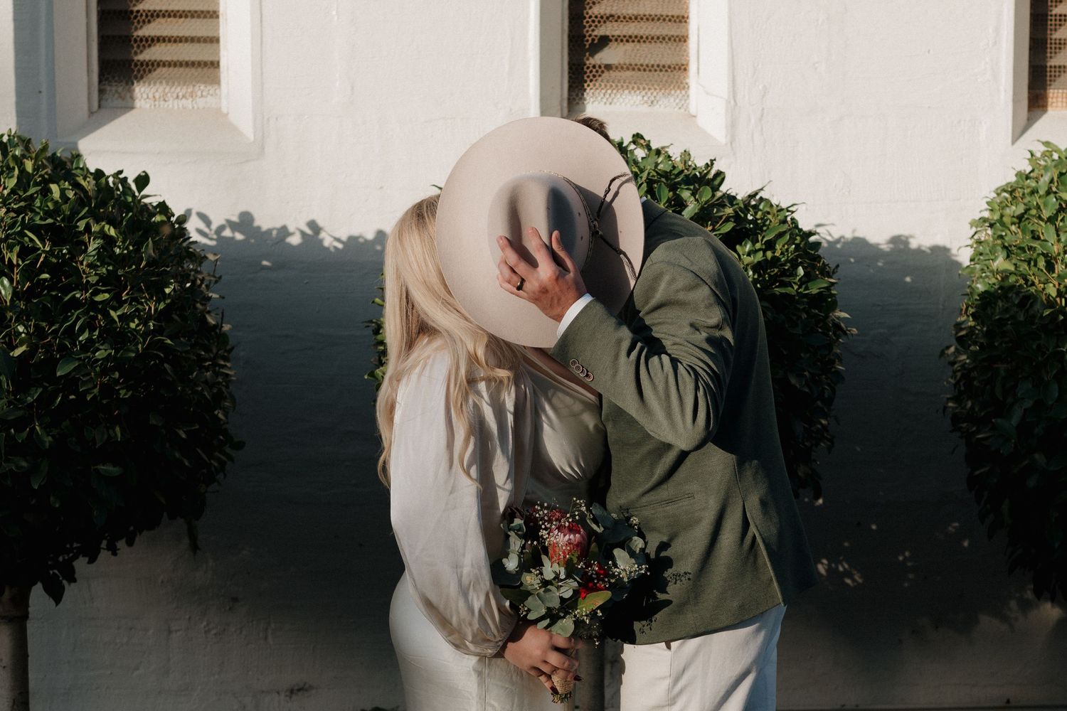 Romantic wedding portraits captured against white building at sunset.