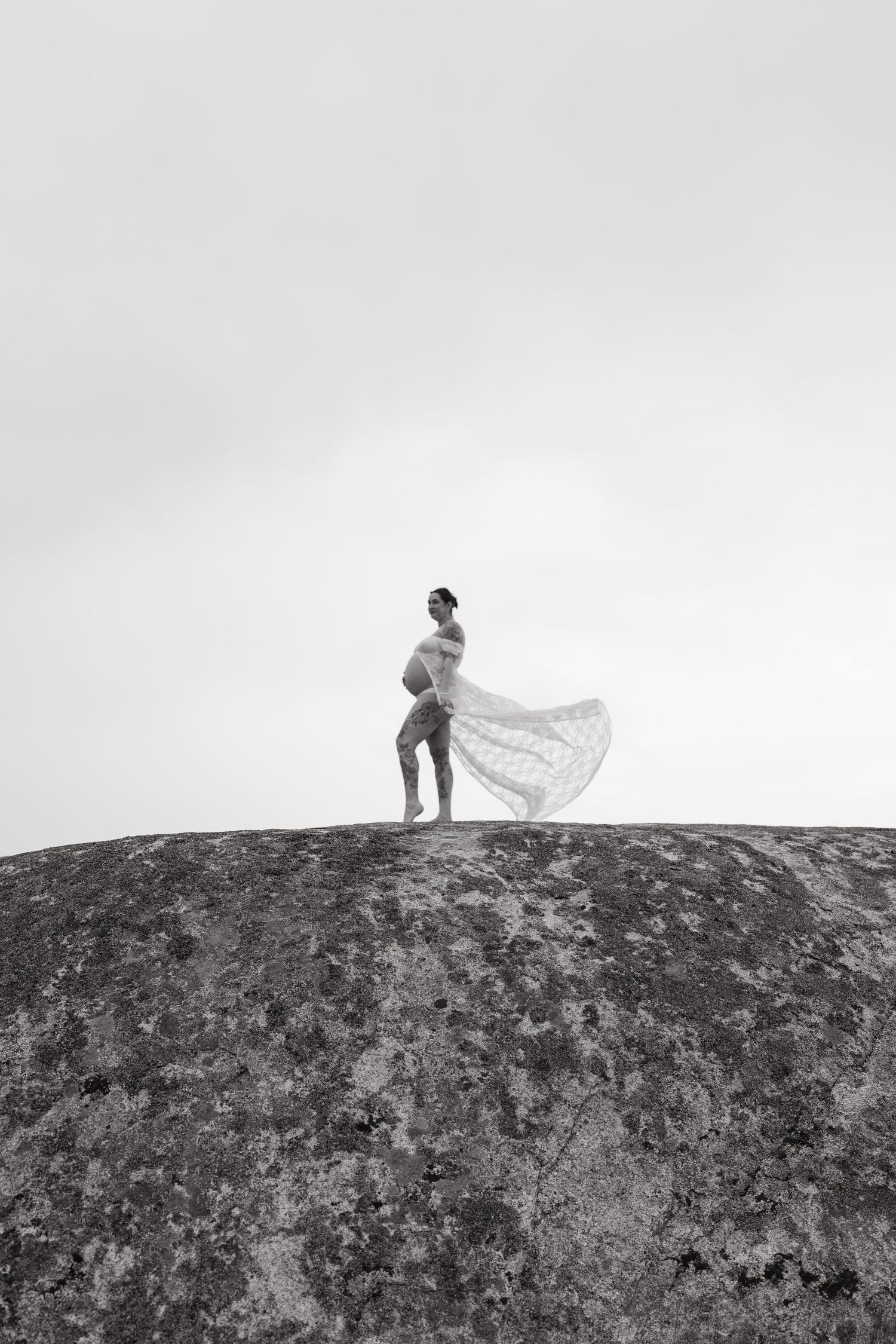 Pregnant woman in flowing lace fabric stands atop a rocky hill in black and white maternity photography.