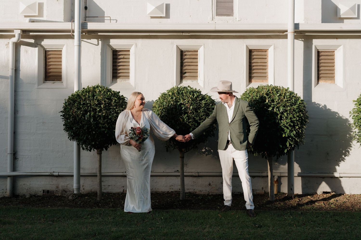 Wedding party members stand in white outfits against a white building exterior.