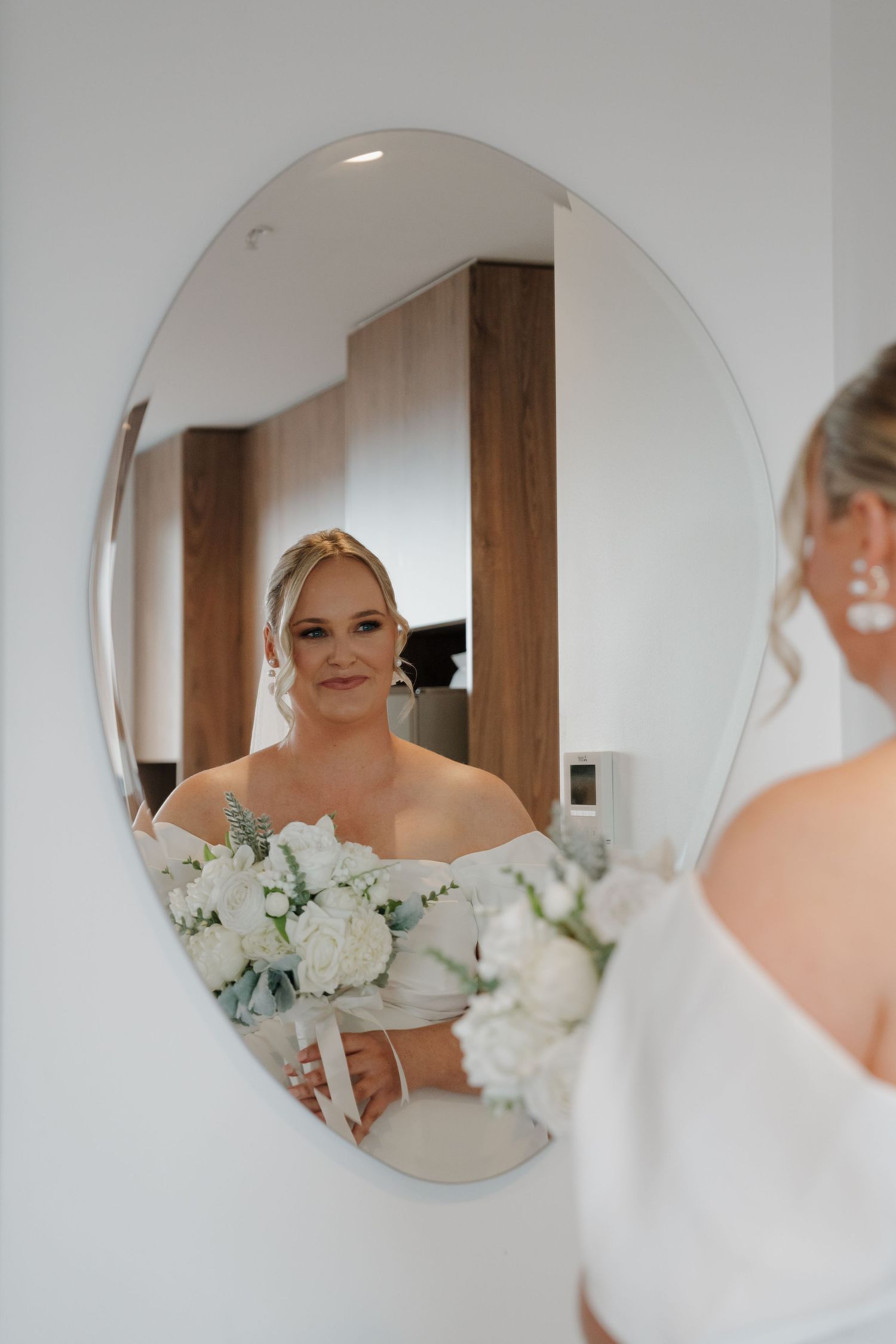 Bride in white off-shoulder gown holding white floral bouquet, admiring her reflection in an oval mirror.