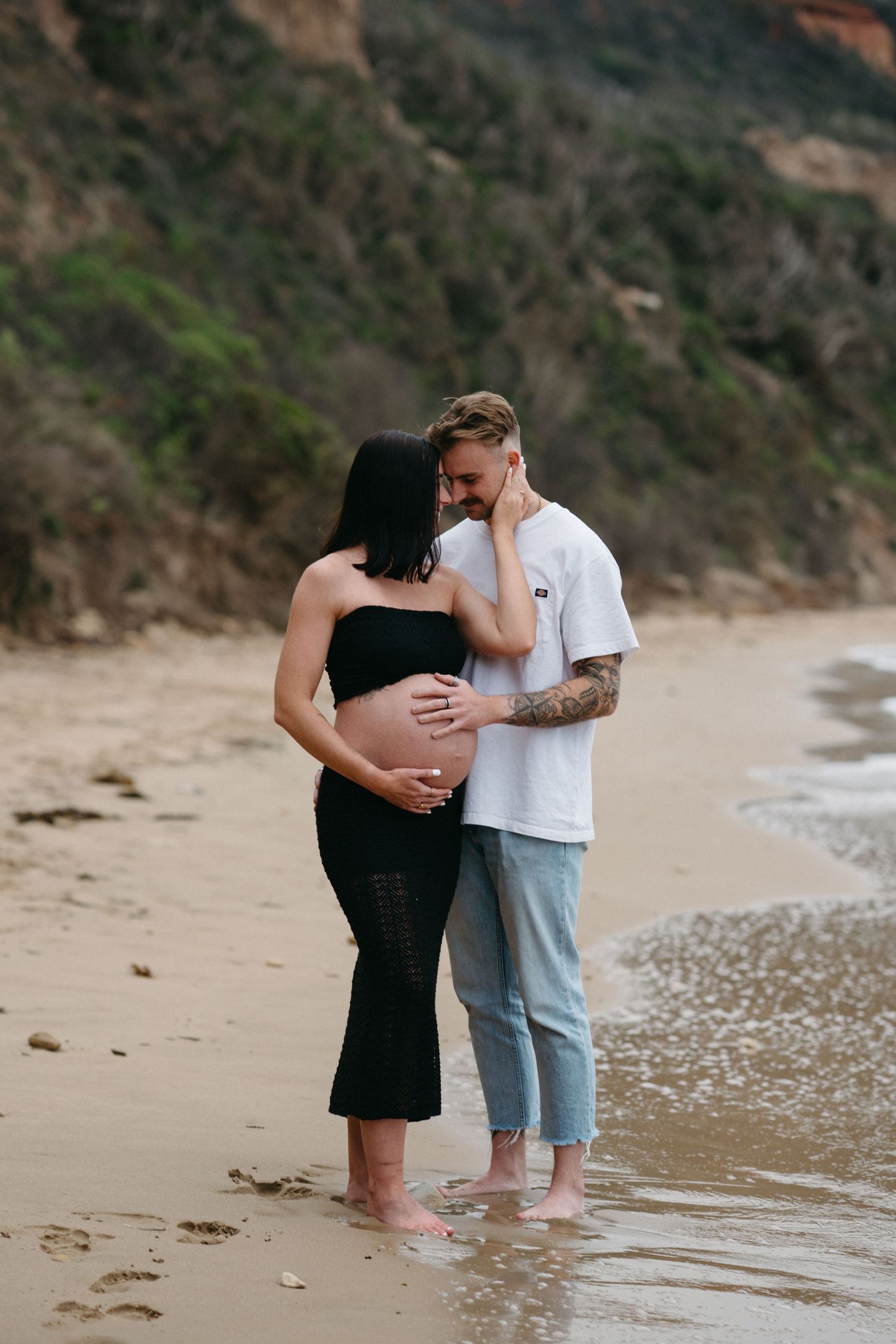 Pregnant couple embracing on sandy beach, woman in black dress cradling baby bump, man in white tee and jeans, coastal cliffs behind.
