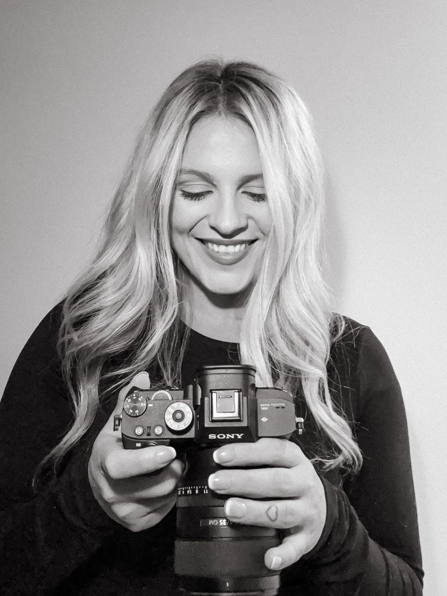 Black and white photo of a smiling blonde woman holding a Sony camera, looking down at the display.