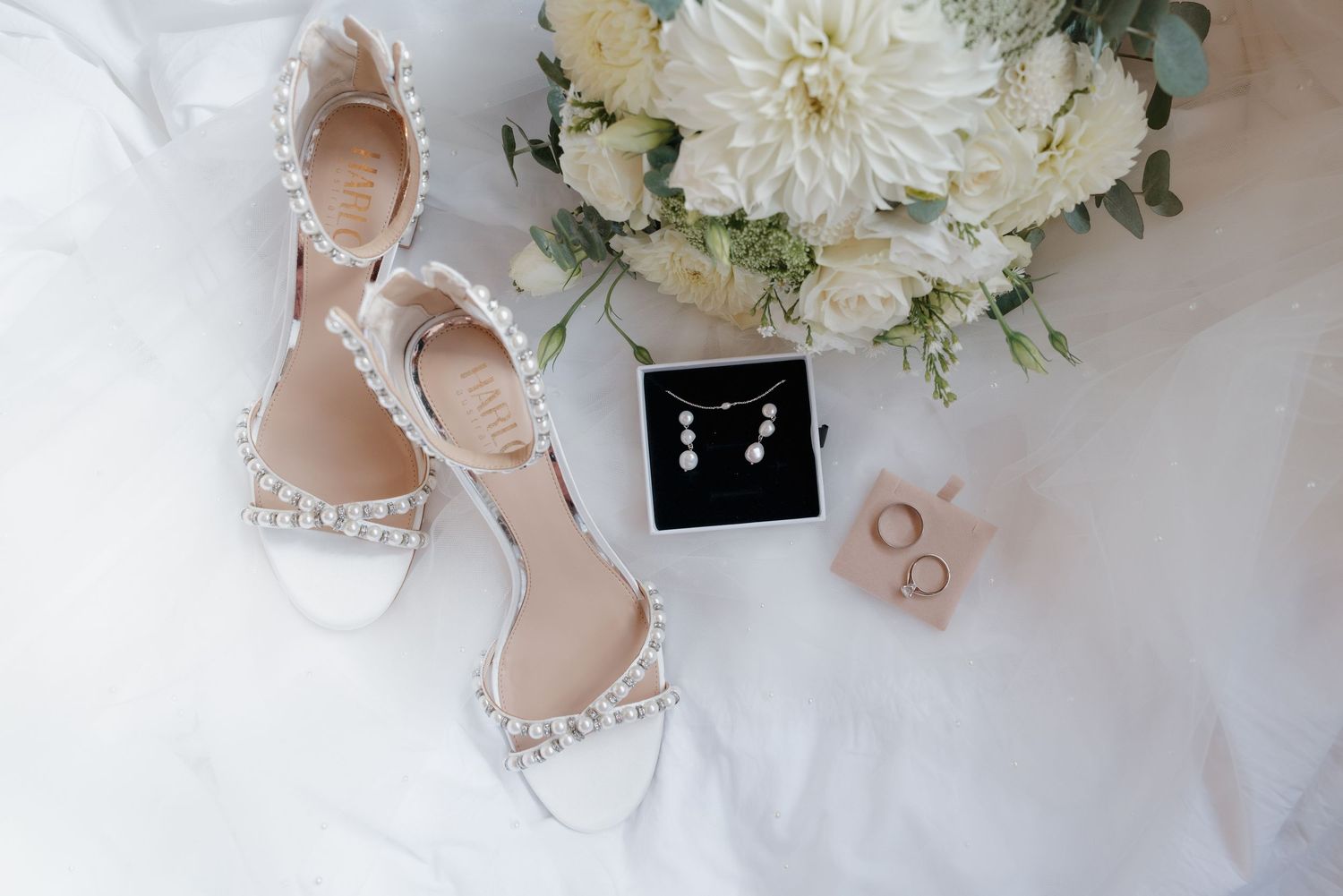 Wedding details flatlay with white flowers, rings, and bridal shoes on light background.