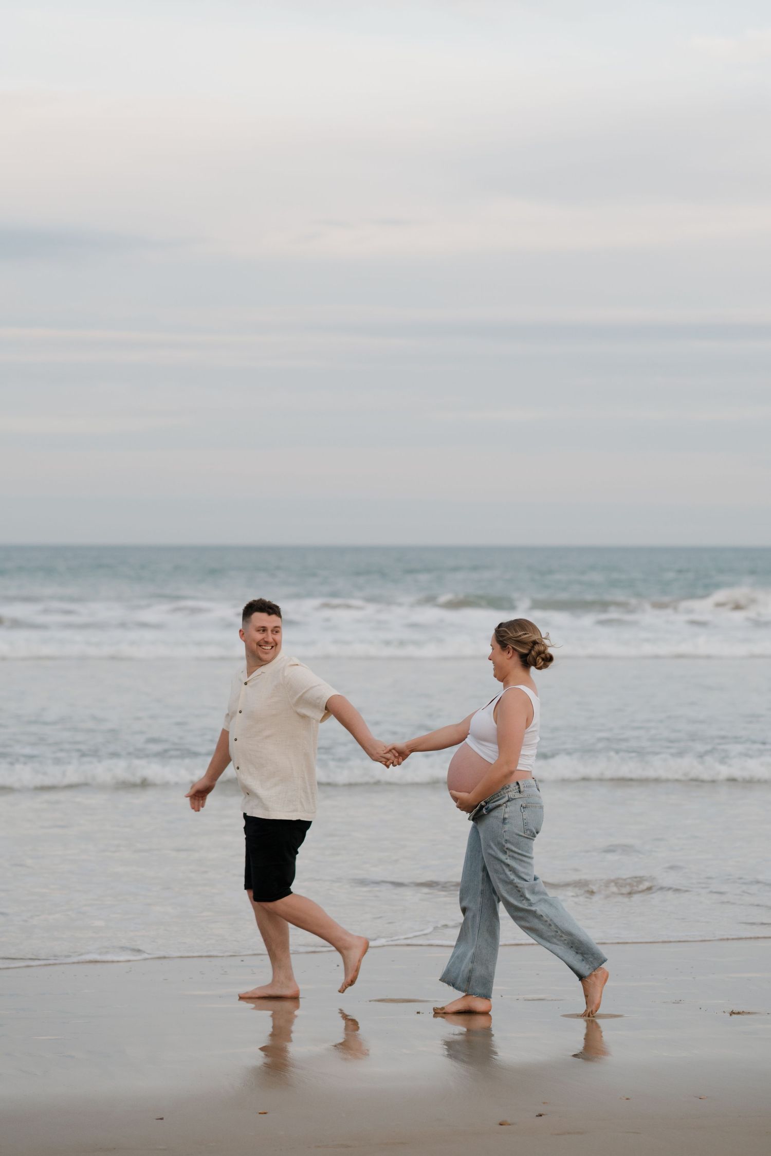 Playful couple holding hands while walking barefoot along the beach shoreline.