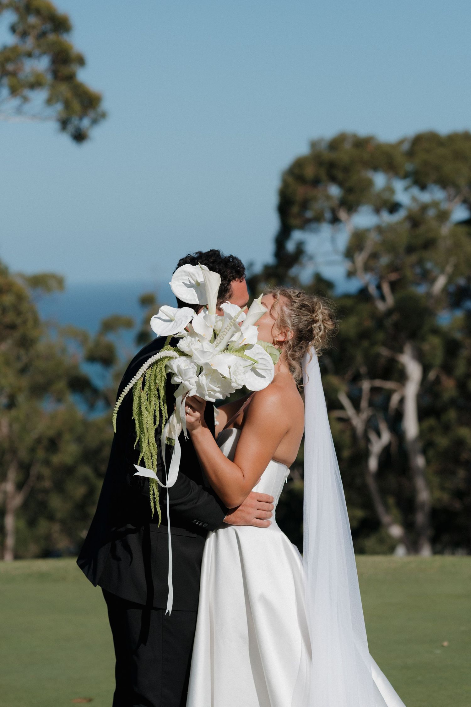 Wedding couple embracing behind large white lilies with ocean view in background.
