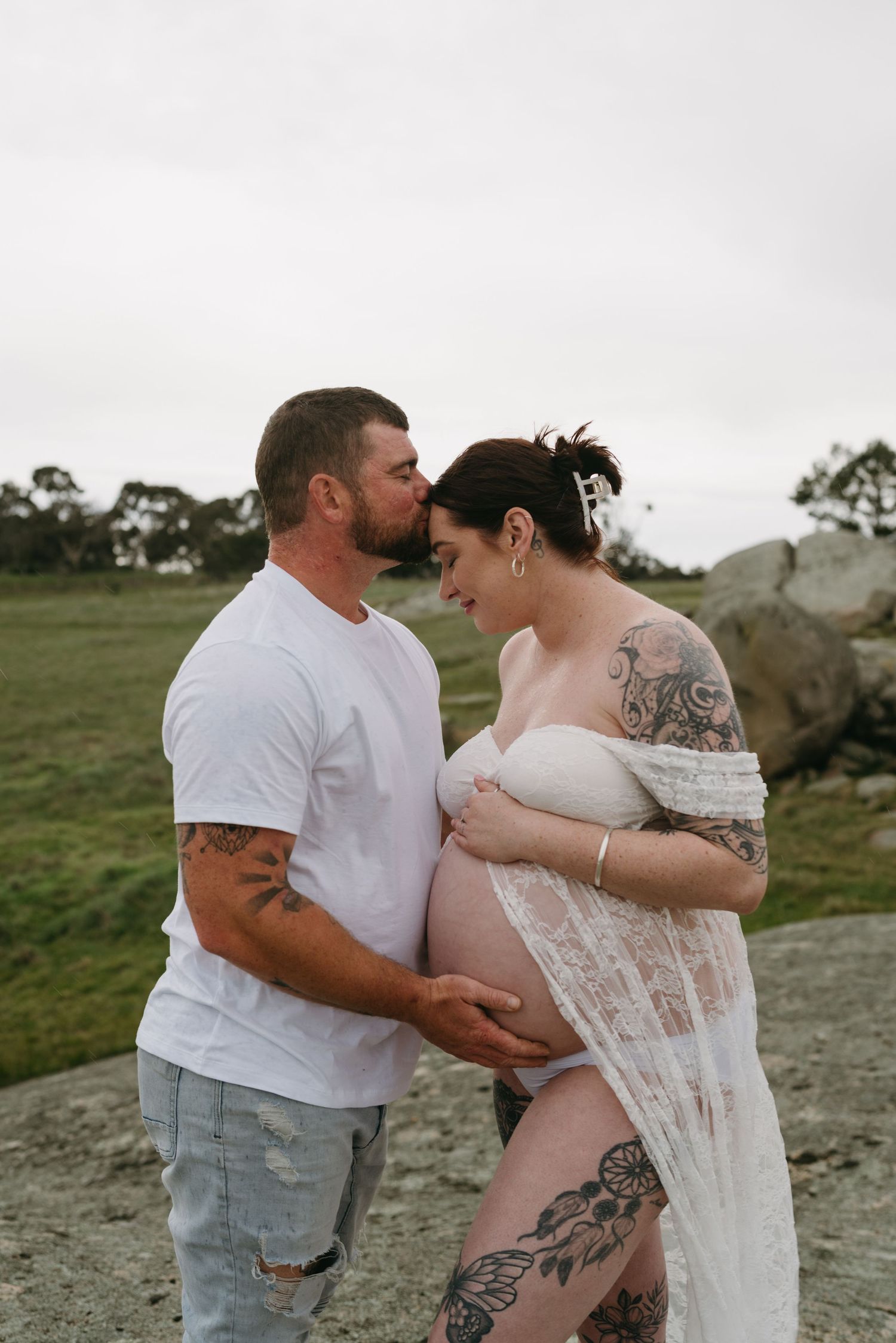 Tattooed couple shares tender maternity photo outdoors, man kissing pregnant woman's head while cradling her baby bump.
