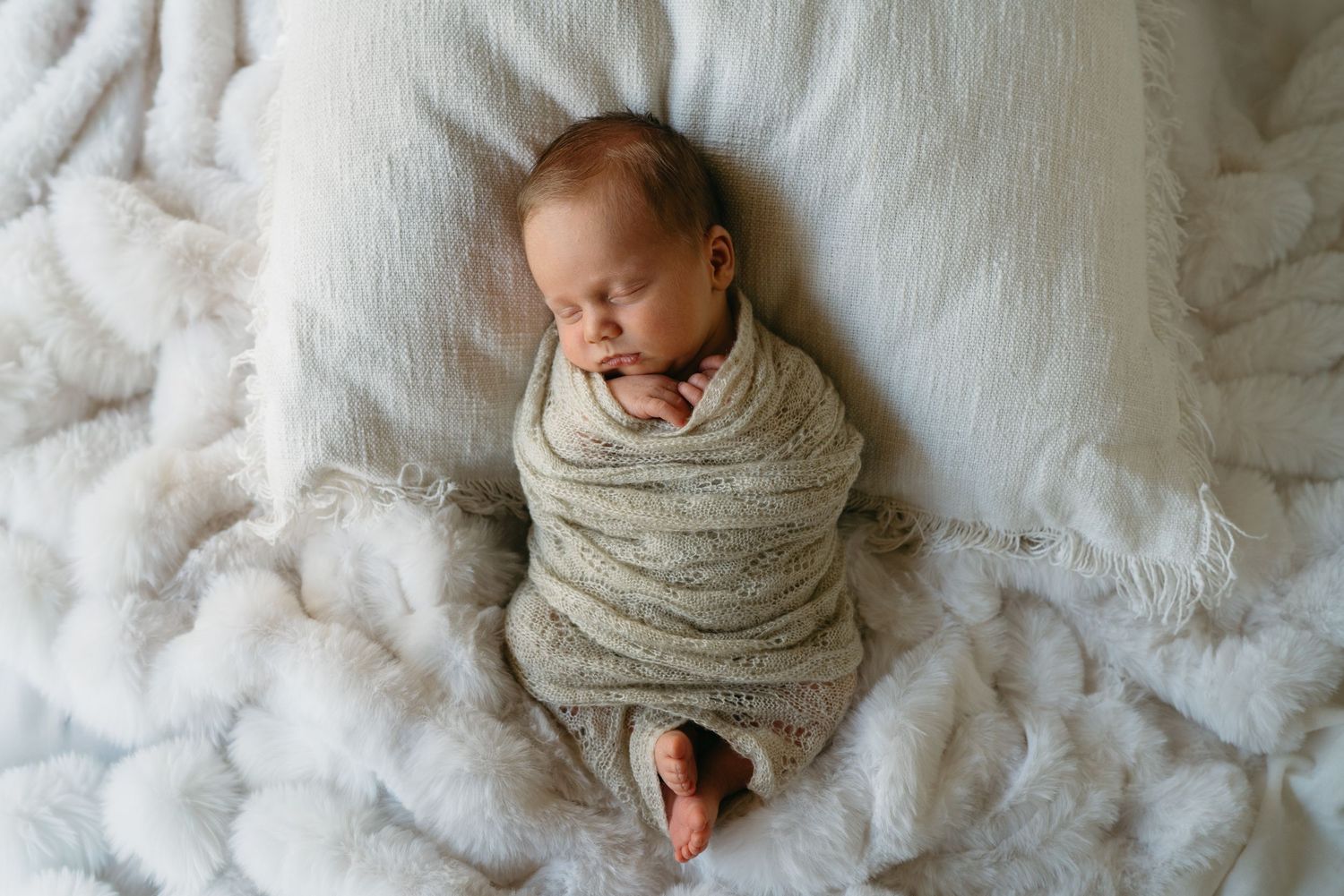Newborn baby swaddled in a neutral knit wrap sleeping peacefully on a soft white fluffy blanket.