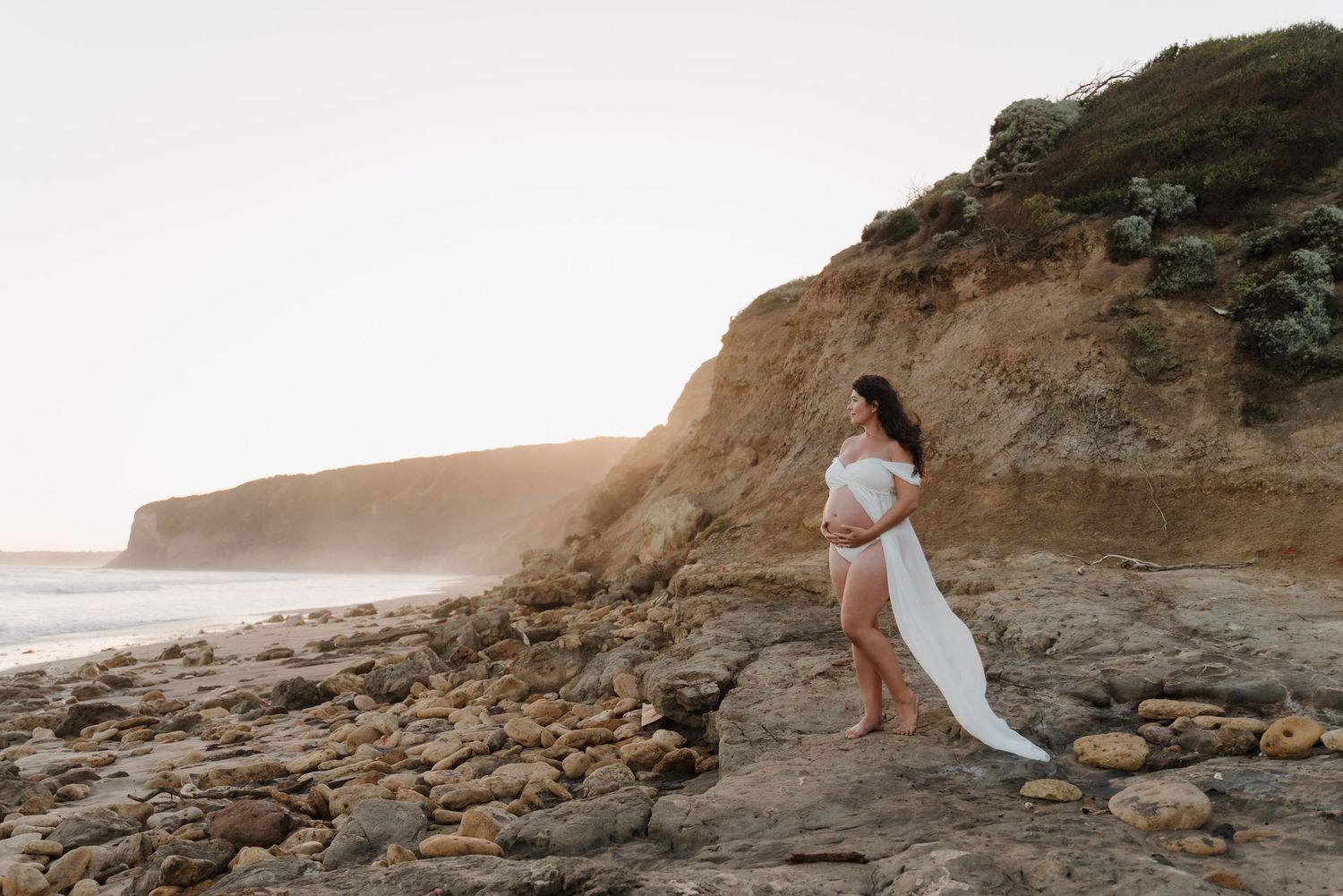 A couple in wedding attire poses romantically on a scenic rocky beach cliff at sunset.