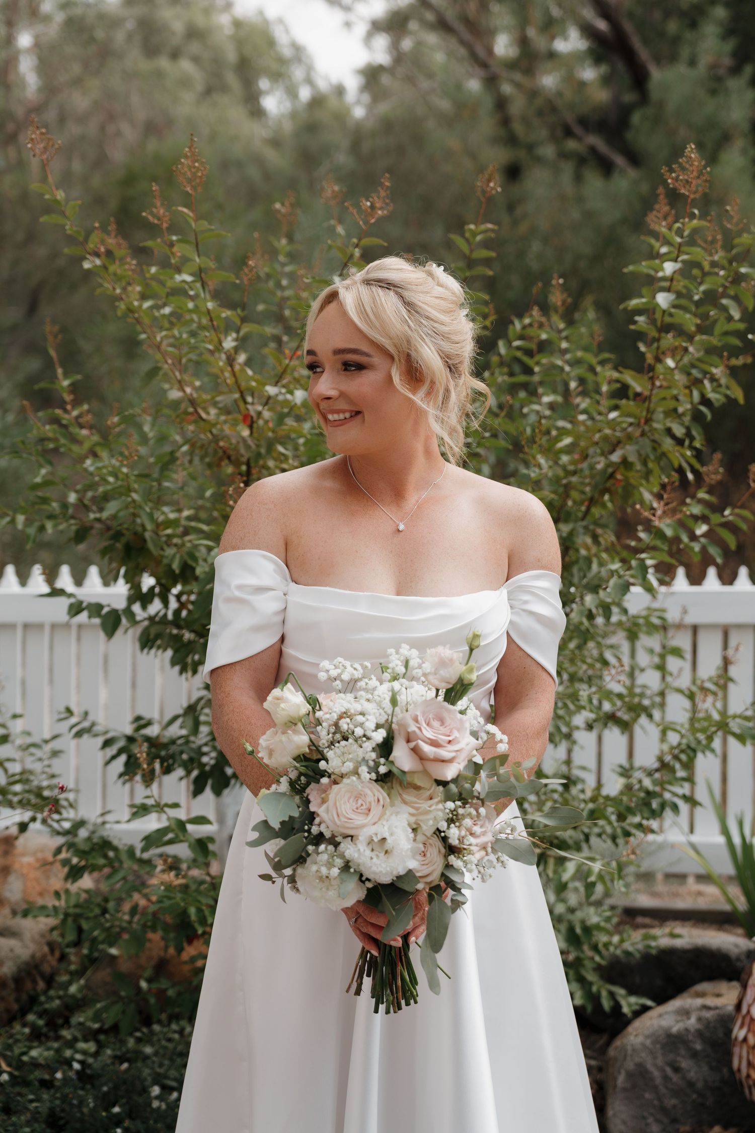 Off-shoulder white wedding dress with delicate bouquet against white picket fence.