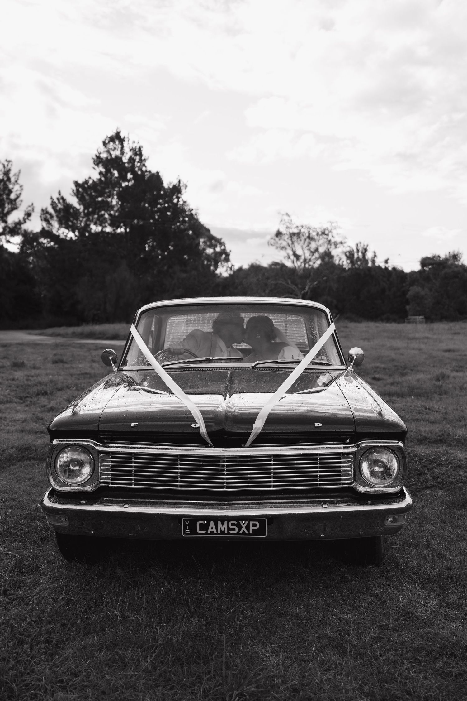 Black and white photo of a vintage Ford car decorated with white ribbons parked on grass, license plate CAMSXP.