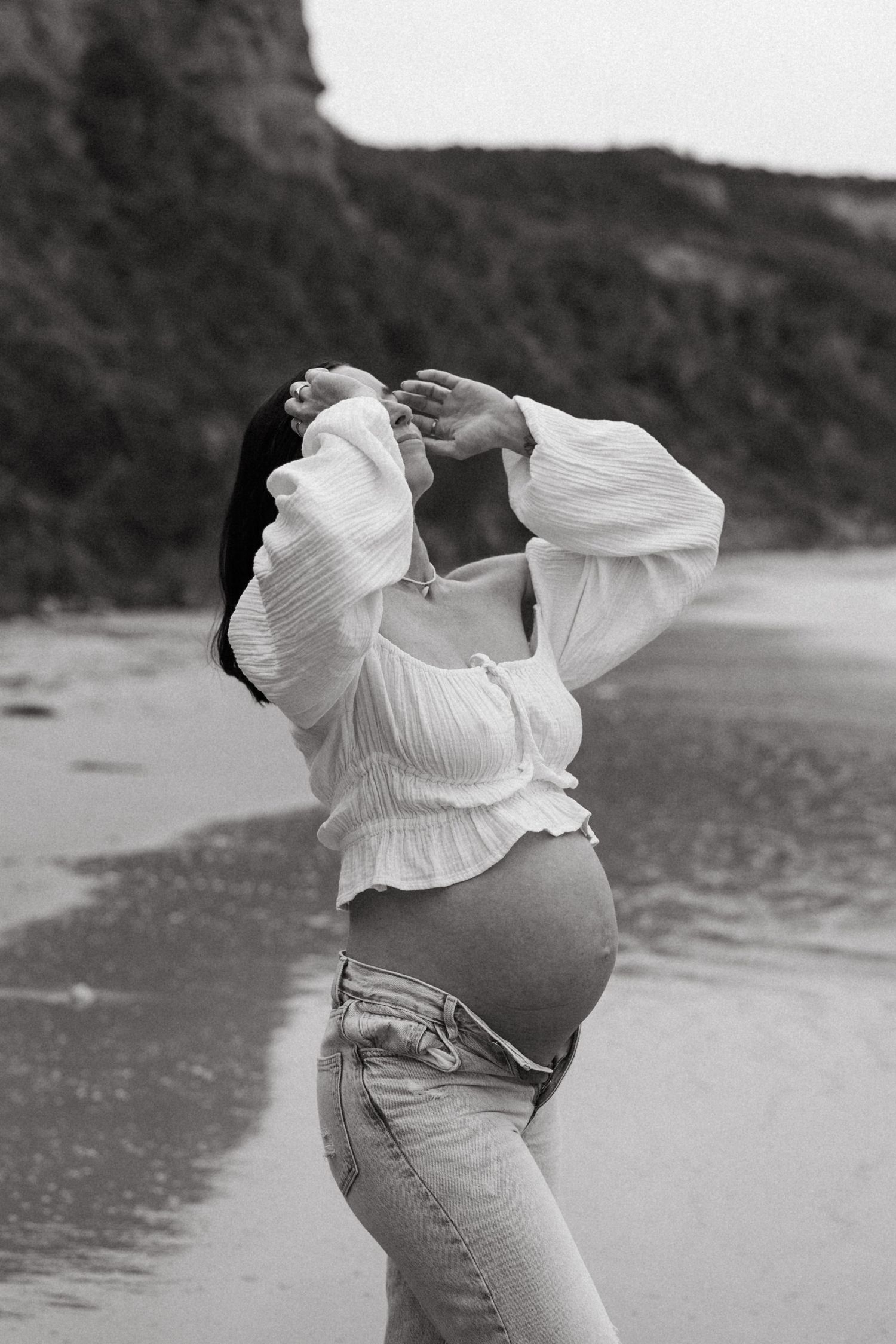Black and white photo of a pregnant woman in a white crop top and jeans posing on a beach with cliffs behind her.