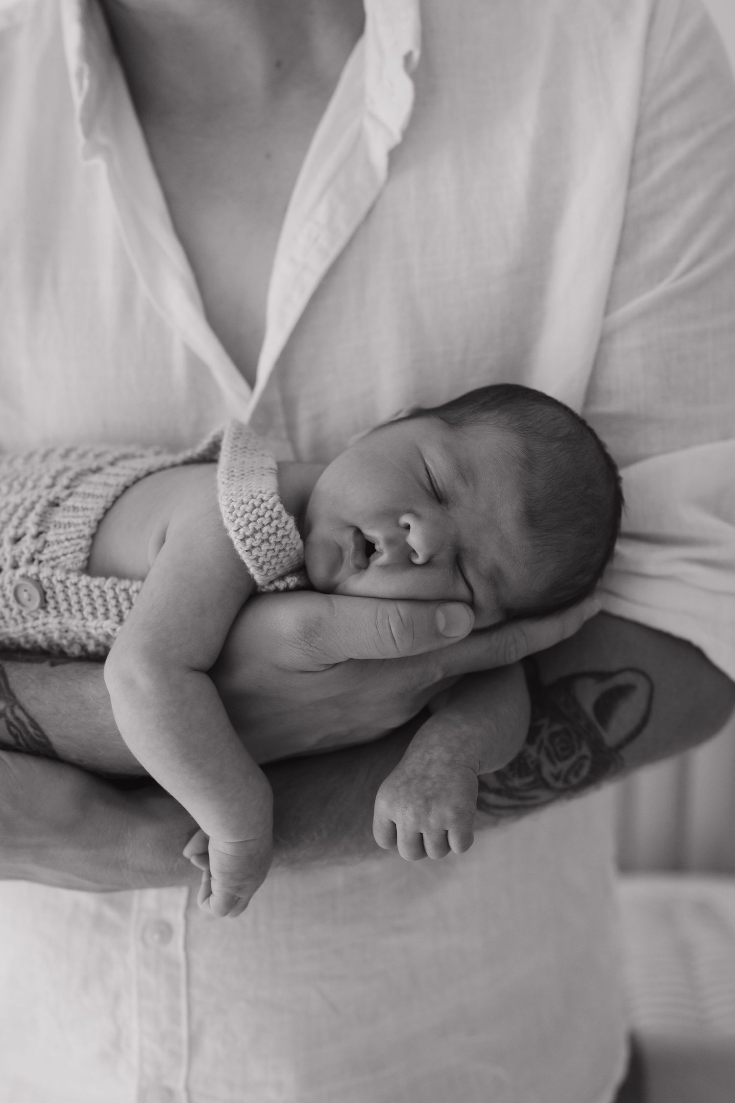 Black and white portrait of sleeping newborn baby cradled in parent's tattooed arms.