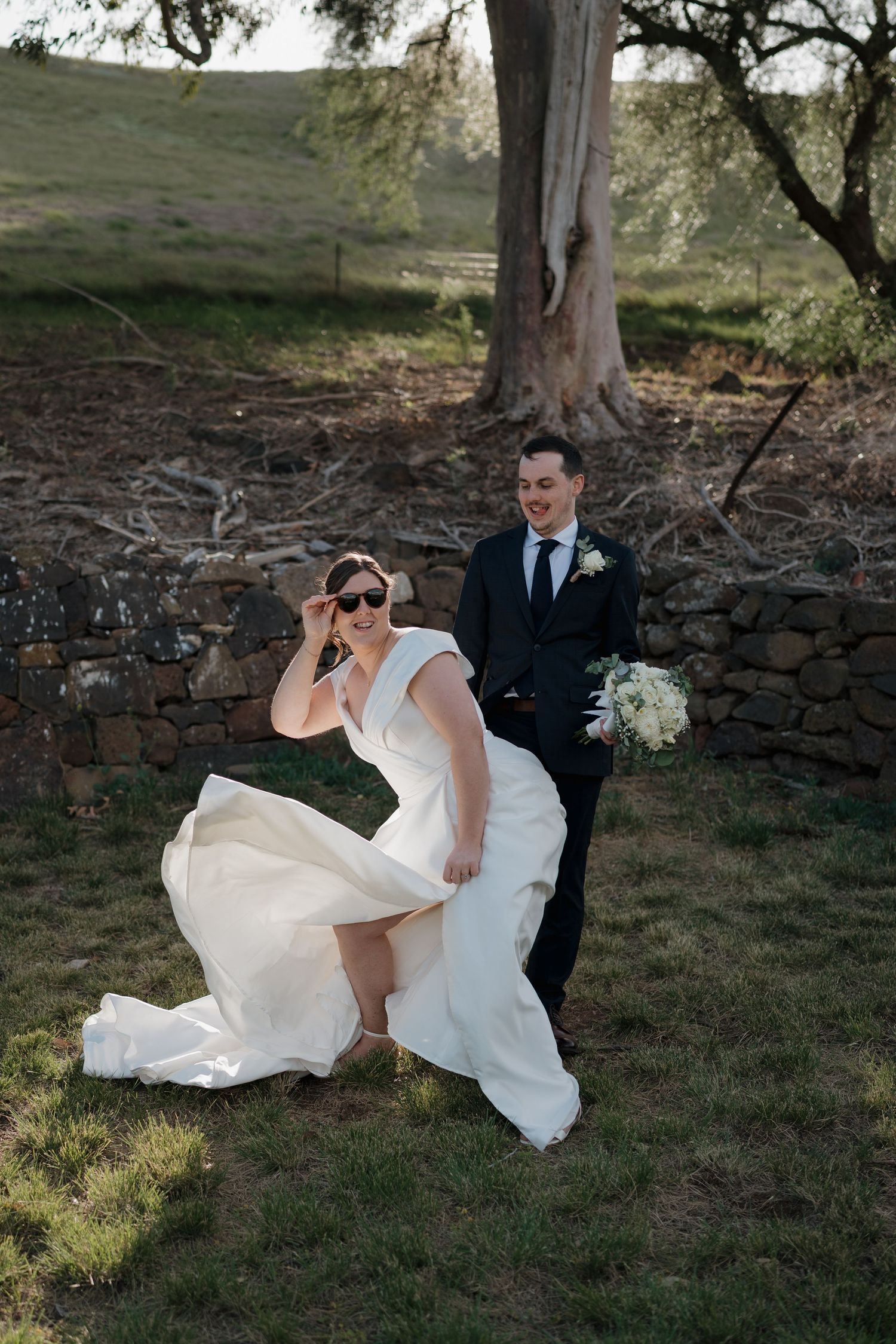 A bride in a flowing white dress and groom share a candid moment outdoors by a rustic stone wall.