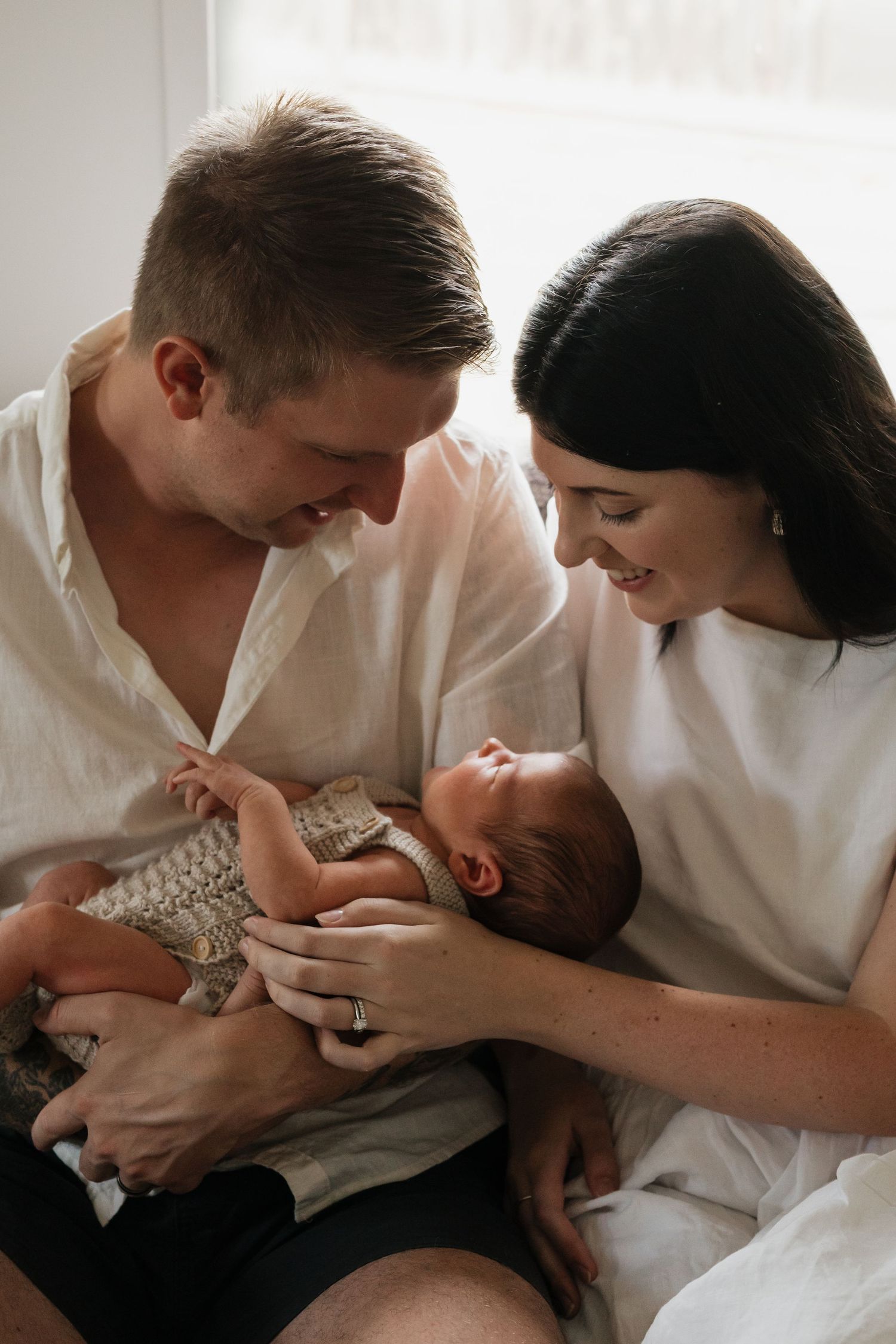 Parents in white shirts sharing loving moments with their infant on a couch.