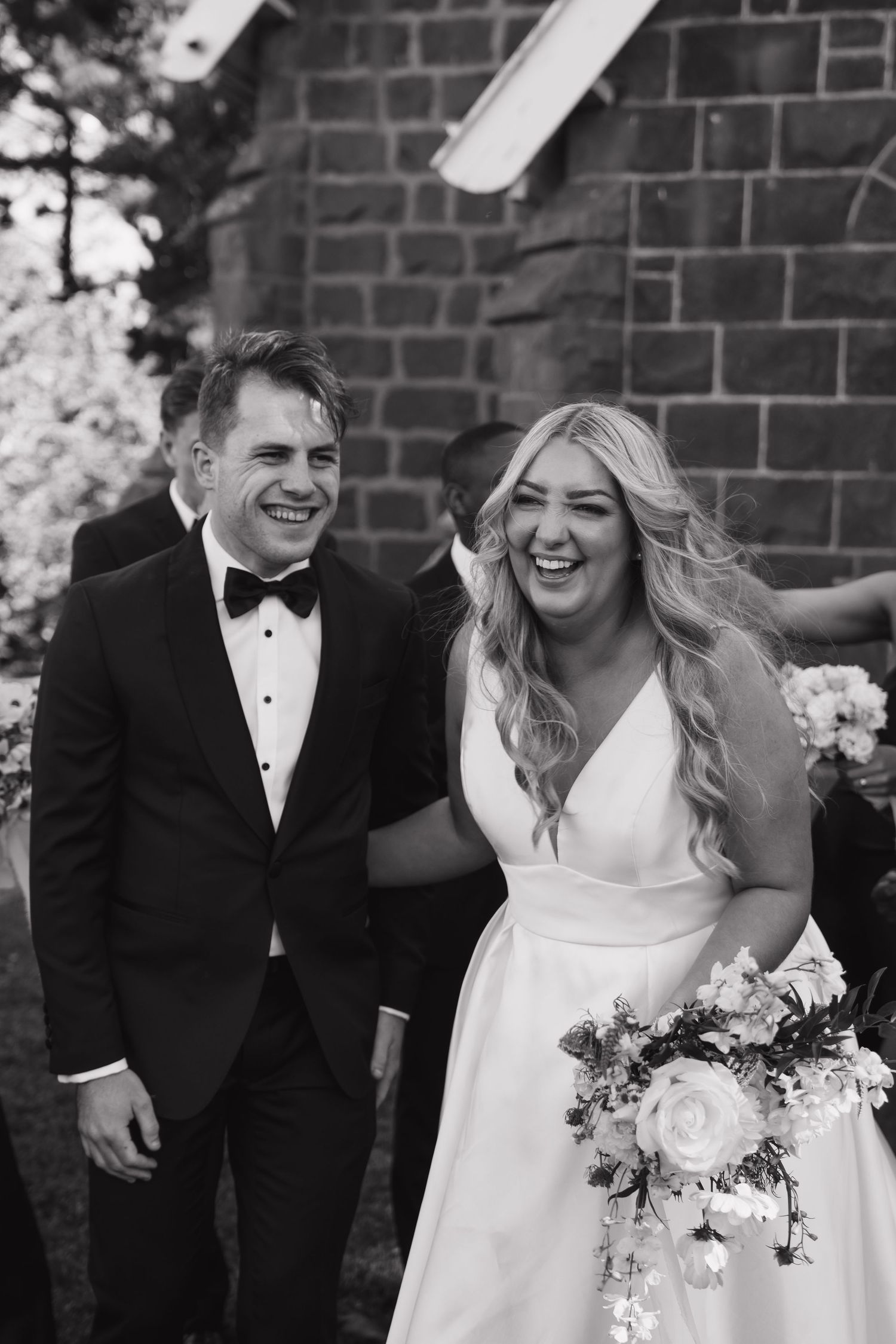 A joyful black and white wedding photo showing a couple laughing together against a brick wall.