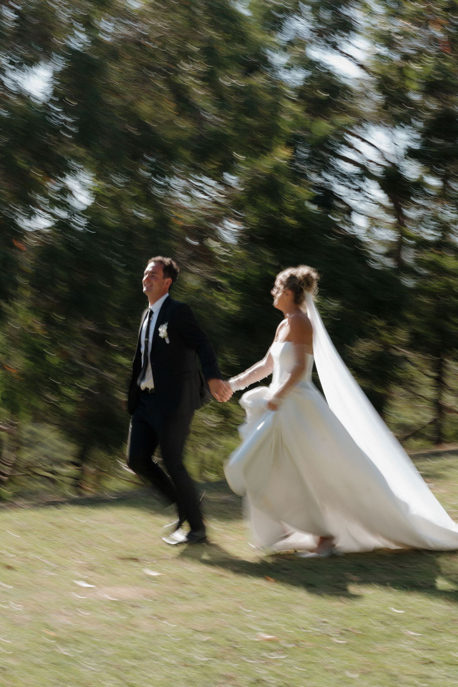 A couple in wedding attire runs playfully through a grassy outdoor setting.