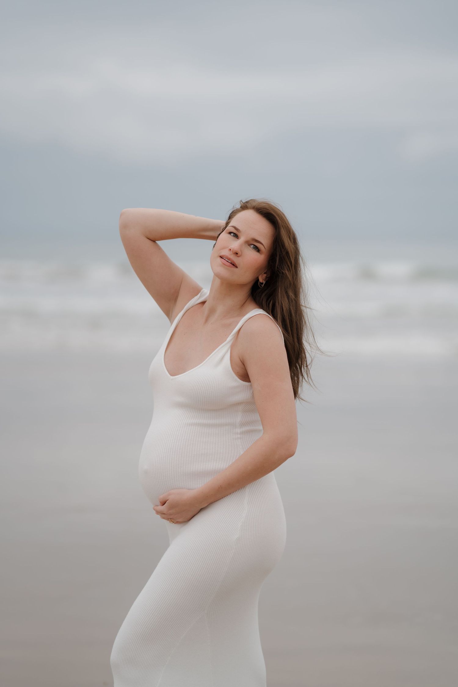 Woman in white dress poses on beach during maternity photoshoot.