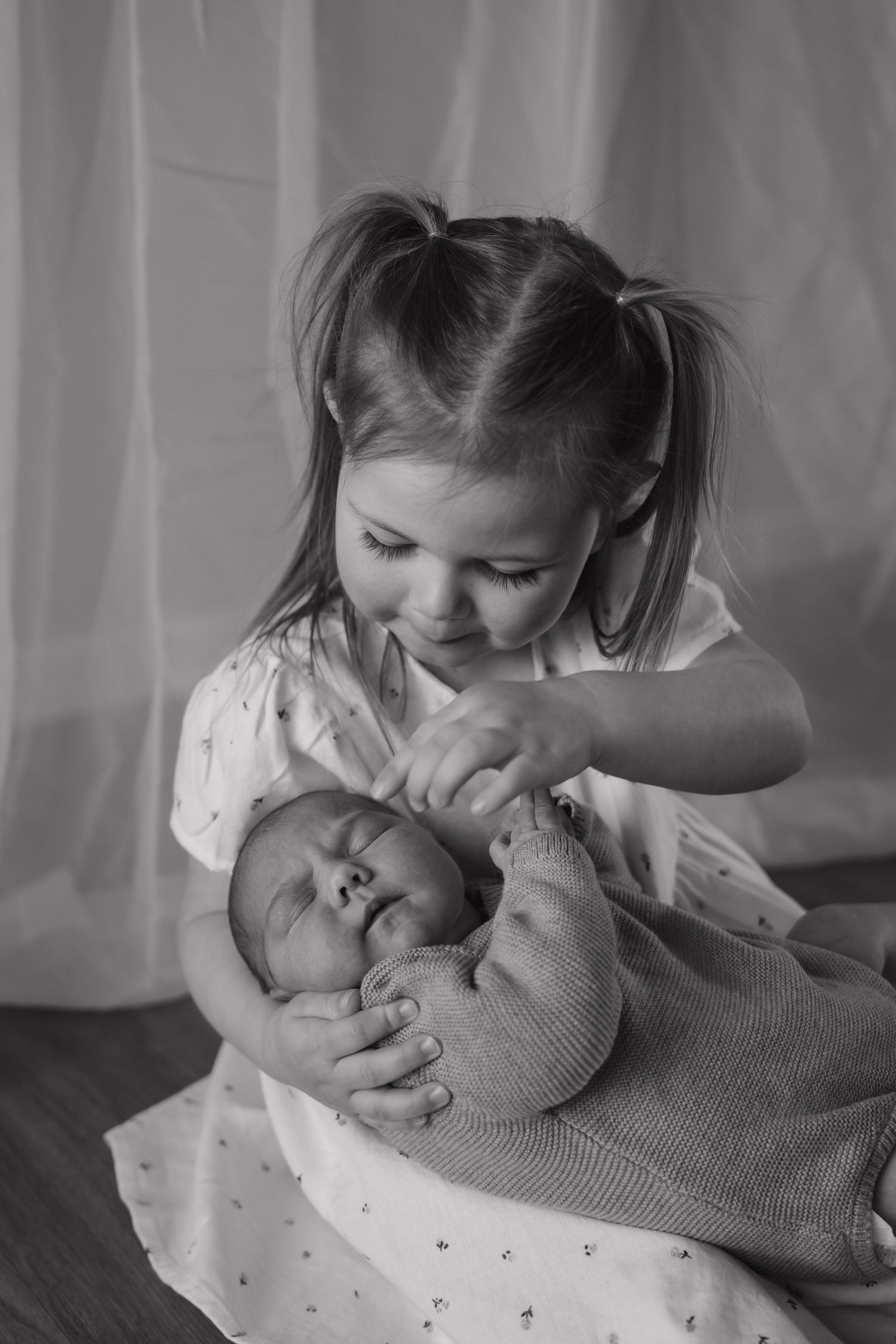 A young child tenderly holds and looks down at a sleeping newborn baby wrapped in a gray blanket.