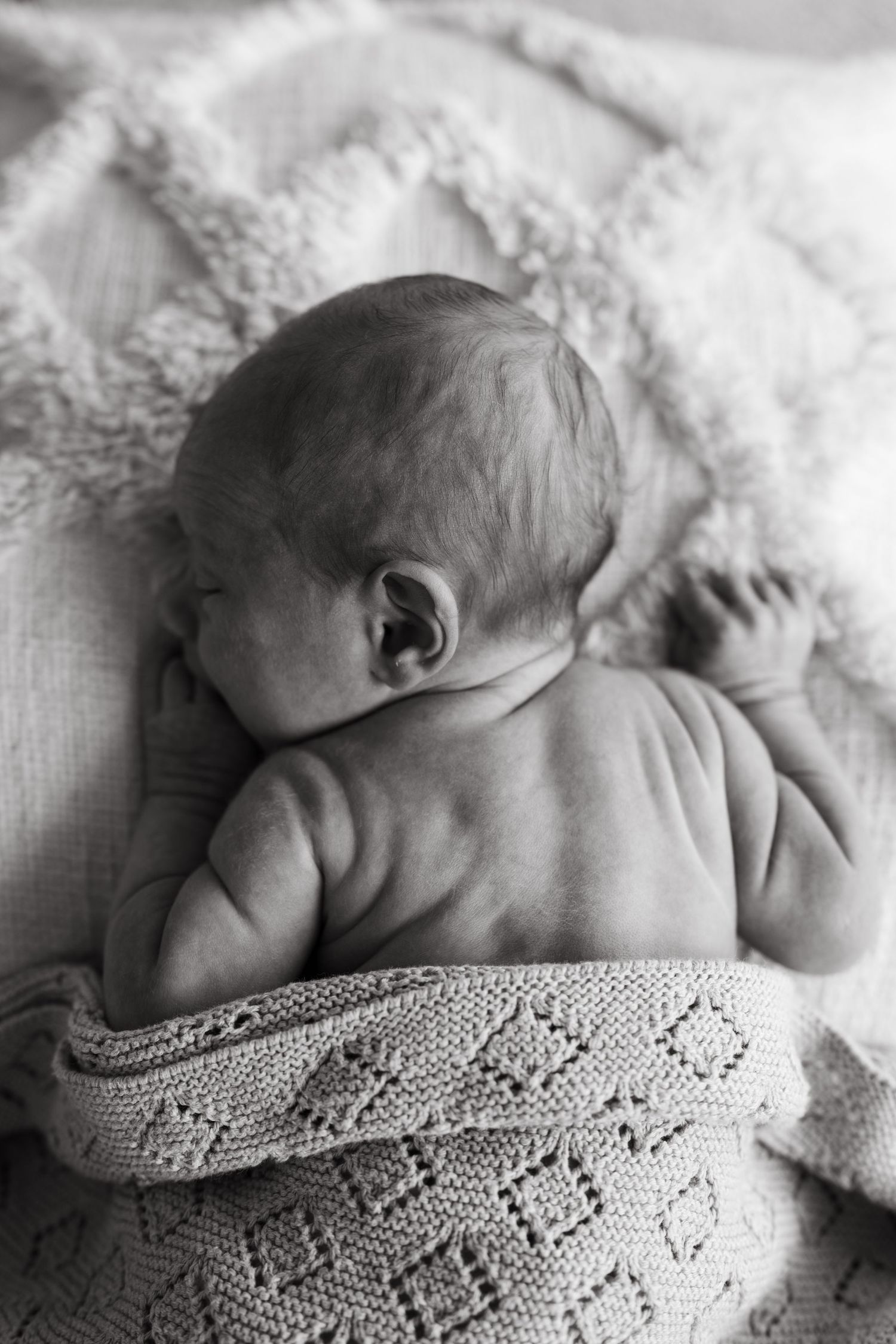 Black and white photo of a sleeping newborn baby wrapped in a knit blanket, viewed from behind.