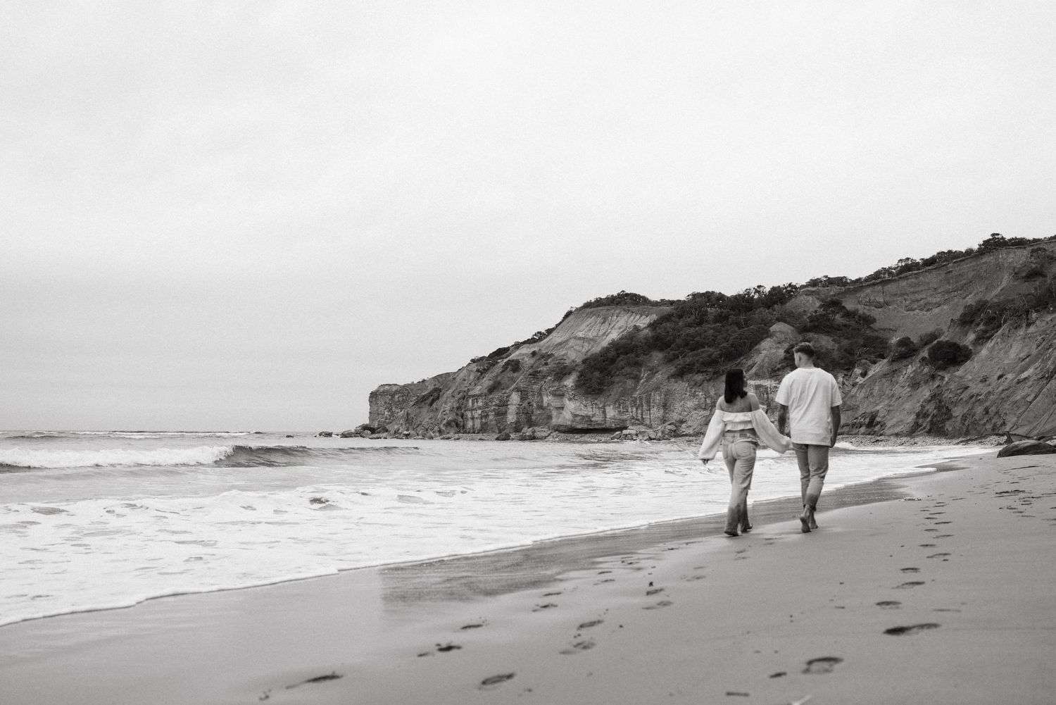 Couple walking hand in hand along a scenic beach with ocean waves and rocky cliffs in black and white.