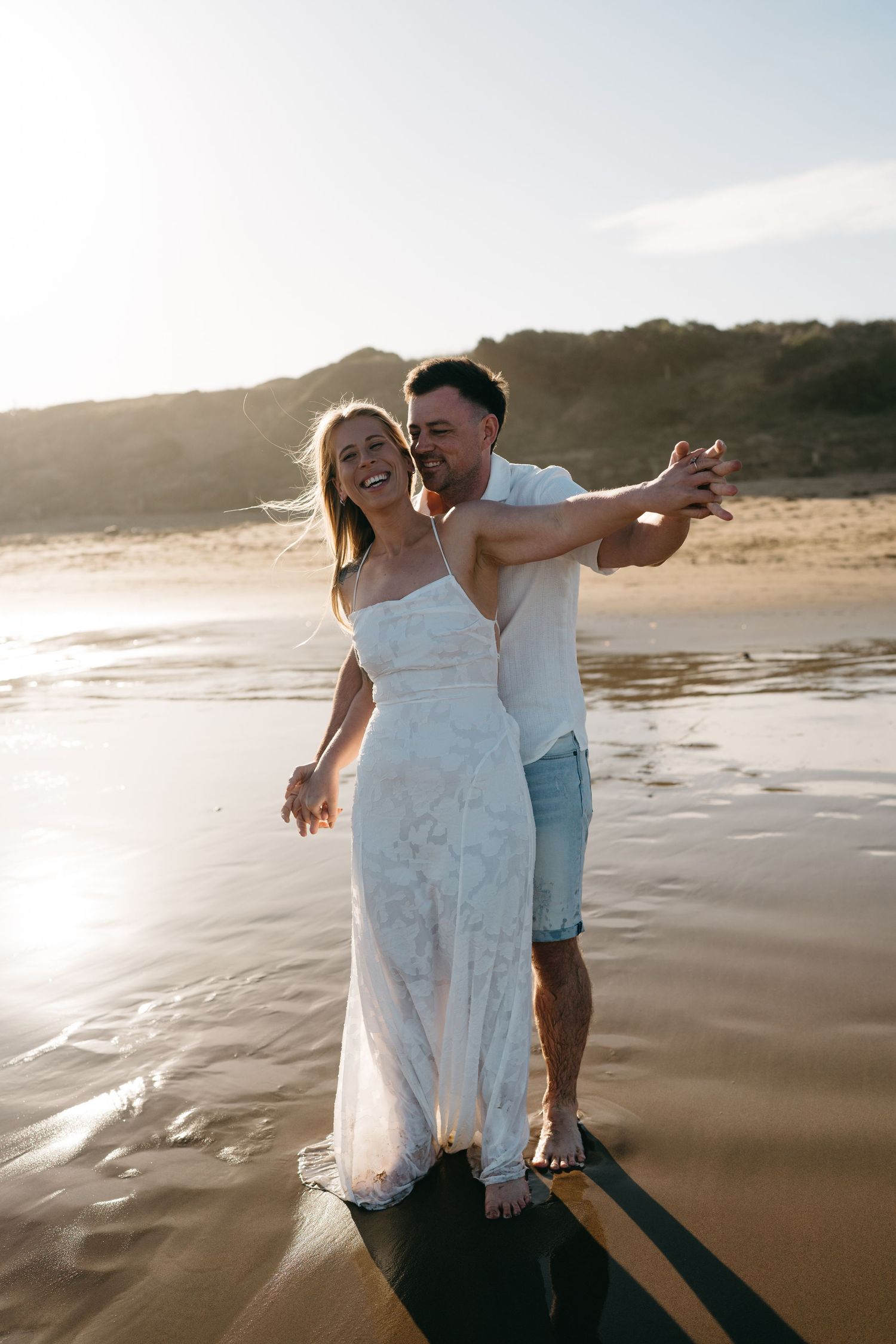 A couple in wedding attire laughing on a beach at golden hour, standing on a surfboard by the ocean.
