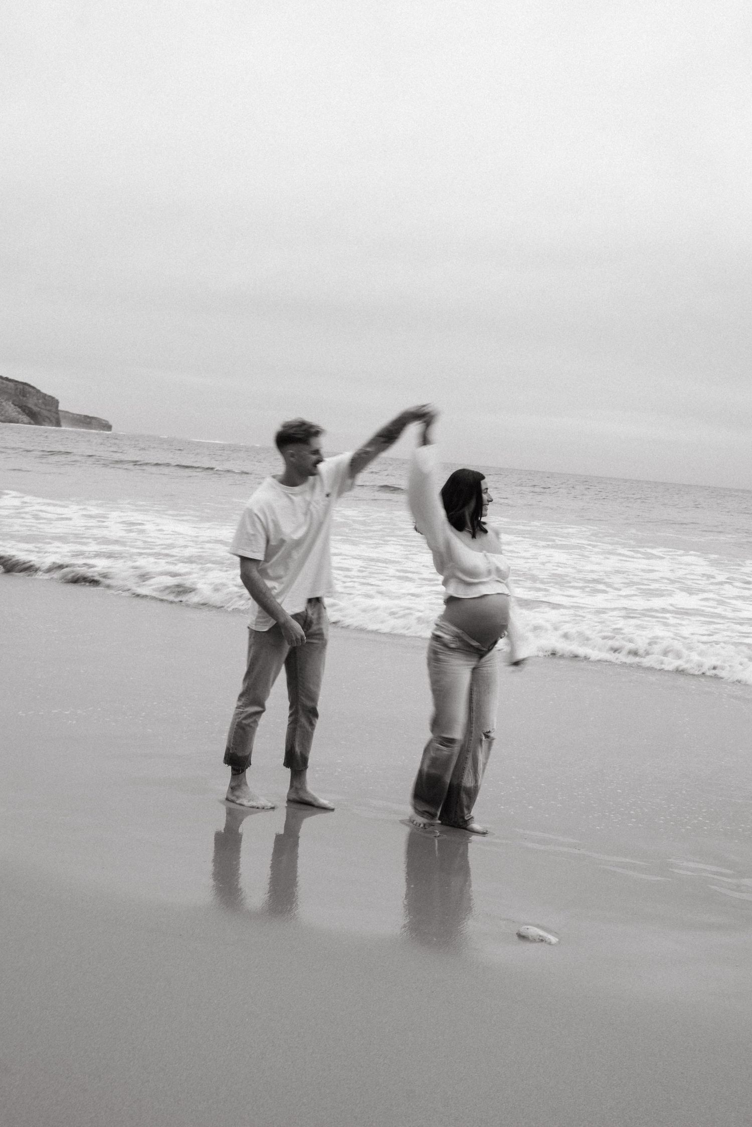 Black and white photo of a couple dancing barefoot on a beach, with ocean waves in the background.