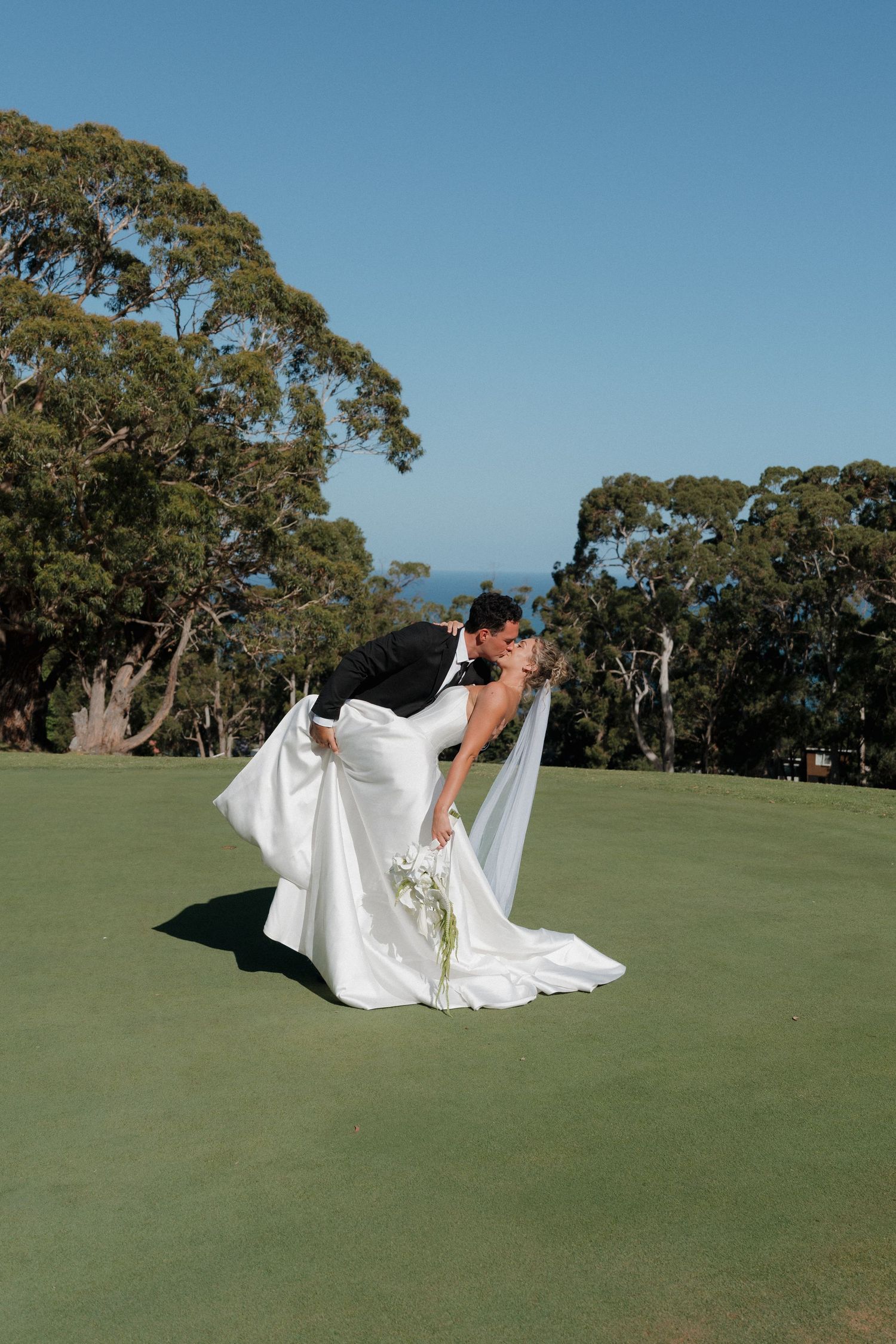 Wedding couple poses dramatically on a green golf course with eucalyptus trees in the background.