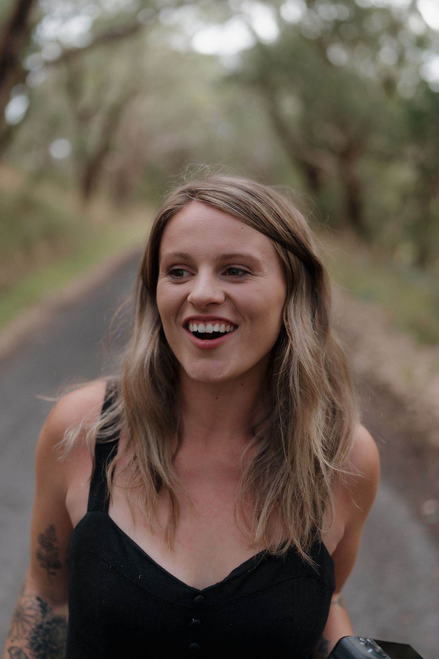 Smiling young woman with blonde hair and tattoo wearing black top on a tree-lined path outdoors.