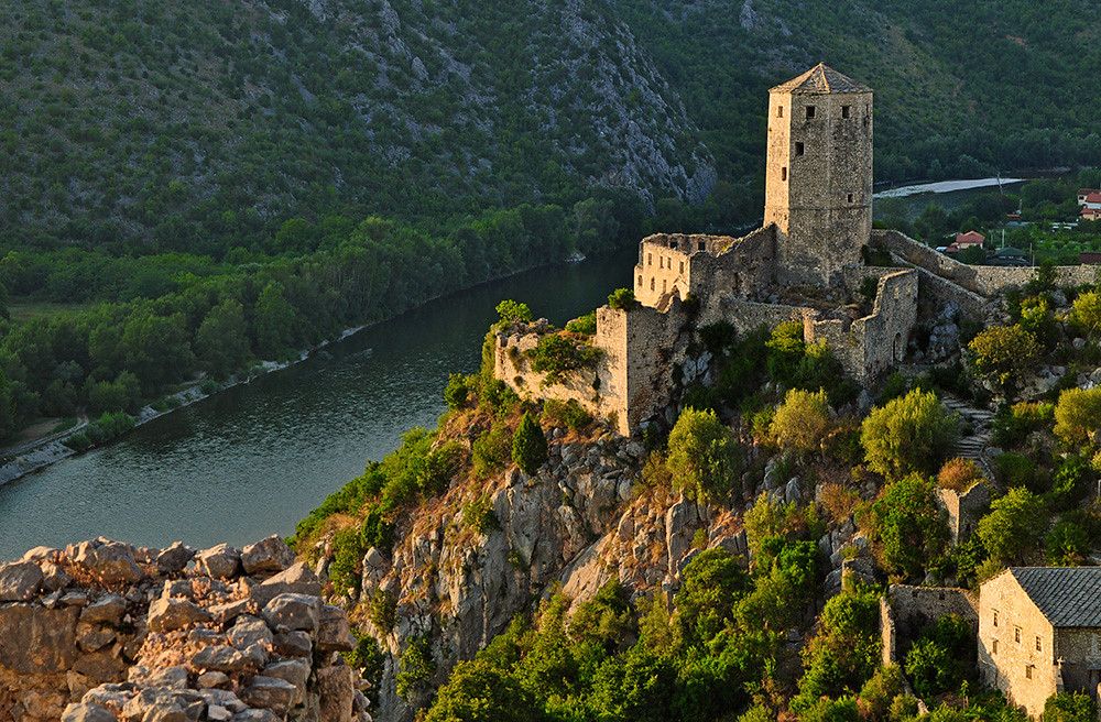 Ancient stone fortress perched dramatically on rocky cliffs overlooking river valley at sunset.