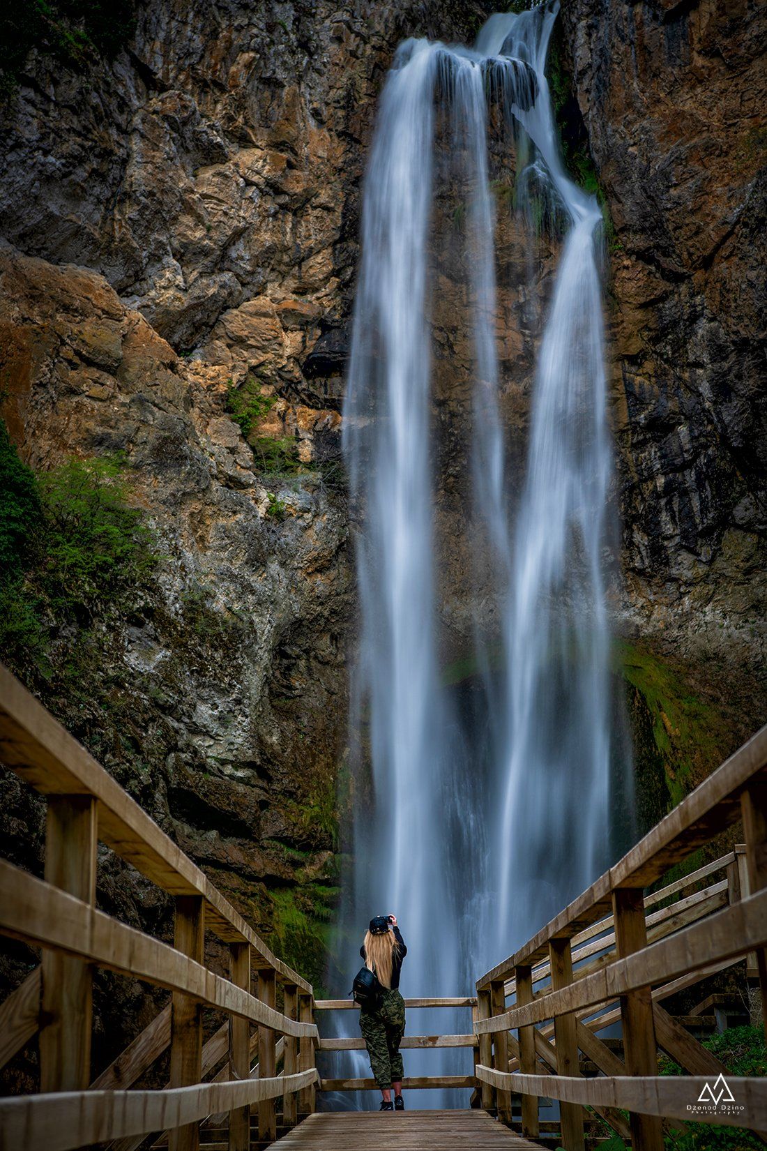 Tall majestic waterfall flowing down rocky cliff face with wooden viewing platform in foreground.