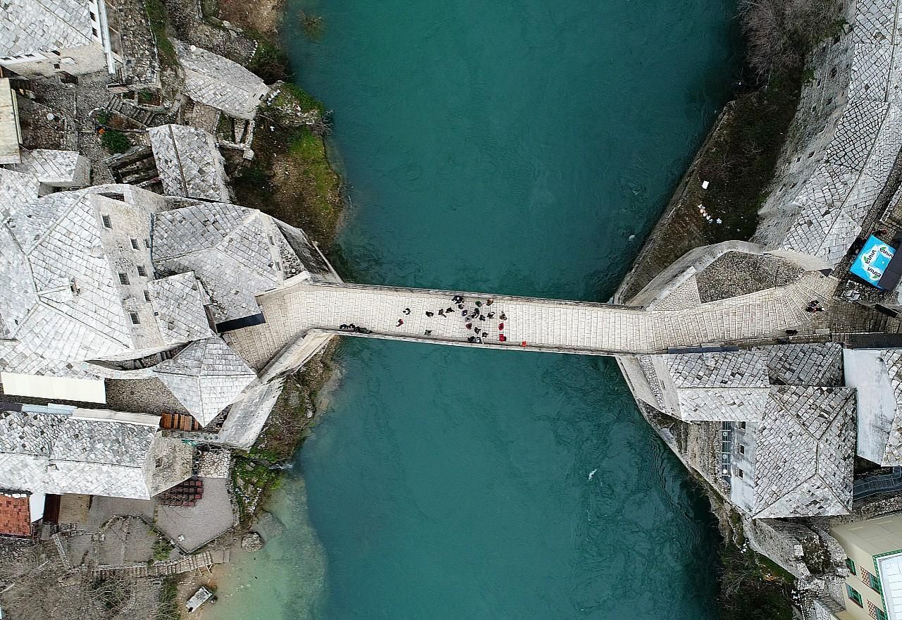 Aerial view of famous Mostar Bridge crossing turquoise river between white stone buildings.